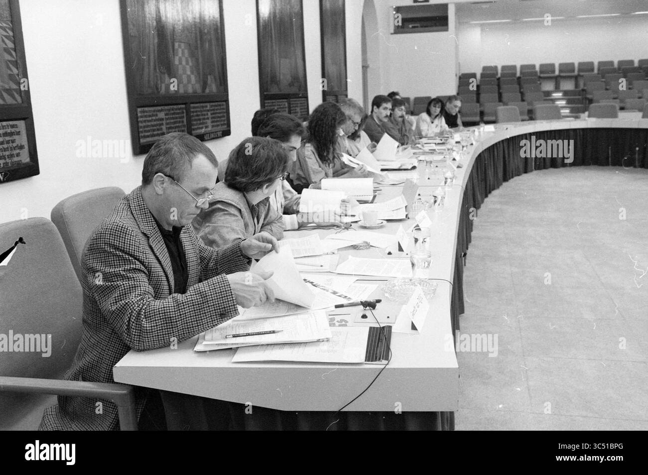 Council Meeting, 00-00-1989 Whizgle News, Dutch Desk, Niederlande, 1950-2000 Eine Gruppe von Einzelpersonen sitzt an einem langen Konferenztisch, wo sie an Diskussionen und der Überprüfung von Dokumenten beteiligt sind. Jeder Teilnehmer scheint sich auf die vor ihm liegenden Materialien konzentriert zu haben, wobei einige Notizen machen und andere die geteilten Informationen in Betracht ziehen. Der Raum ist halbrund gestaltet und an den Wänden werden verschiedene Präsentationen gezeigt. Stockfoto