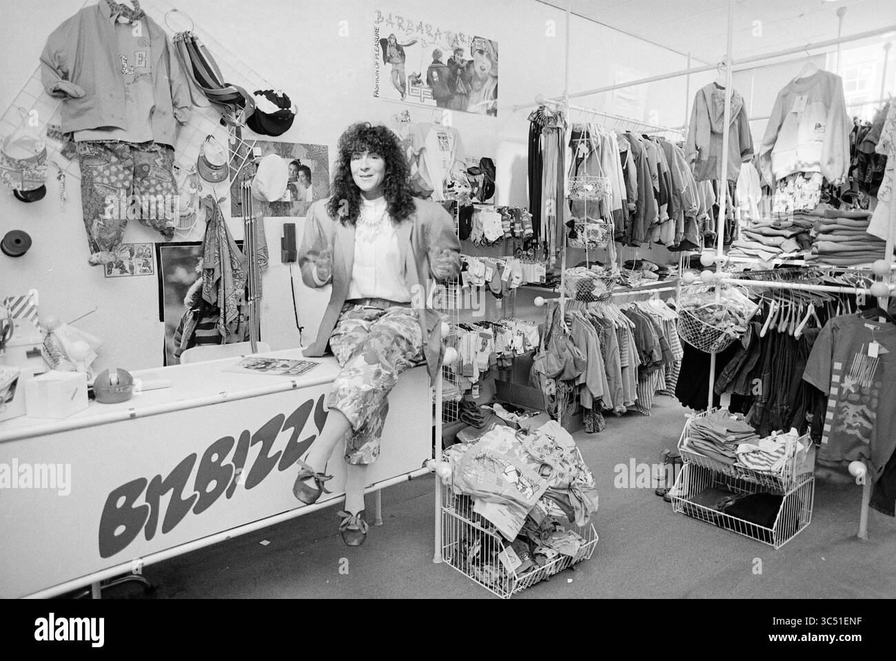 Kindermode Bizbizzy Bloemendaalseweg, Geschäfte und Kioske, Bloemendaal, Bloemendaalseweg, 11-05-1989 Whizgle News, Dutch Desk, Niederlande, 1950-2000 Ein lebhaftes Bekleidungsgeschäft mit einer Vielzahl von farbenfrohen Outfits und Accessoires, in dem ein stilvolles Individuum lässig auf einer Theke sitzt und eine entspannte Atmosphäre bietet. Der Raum ist mit Kleiderregalen in verschiedenen Mustern und Stilen gefüllt, die eine eklektische und einladende Einkaufsumgebung schaffen. Stockfoto