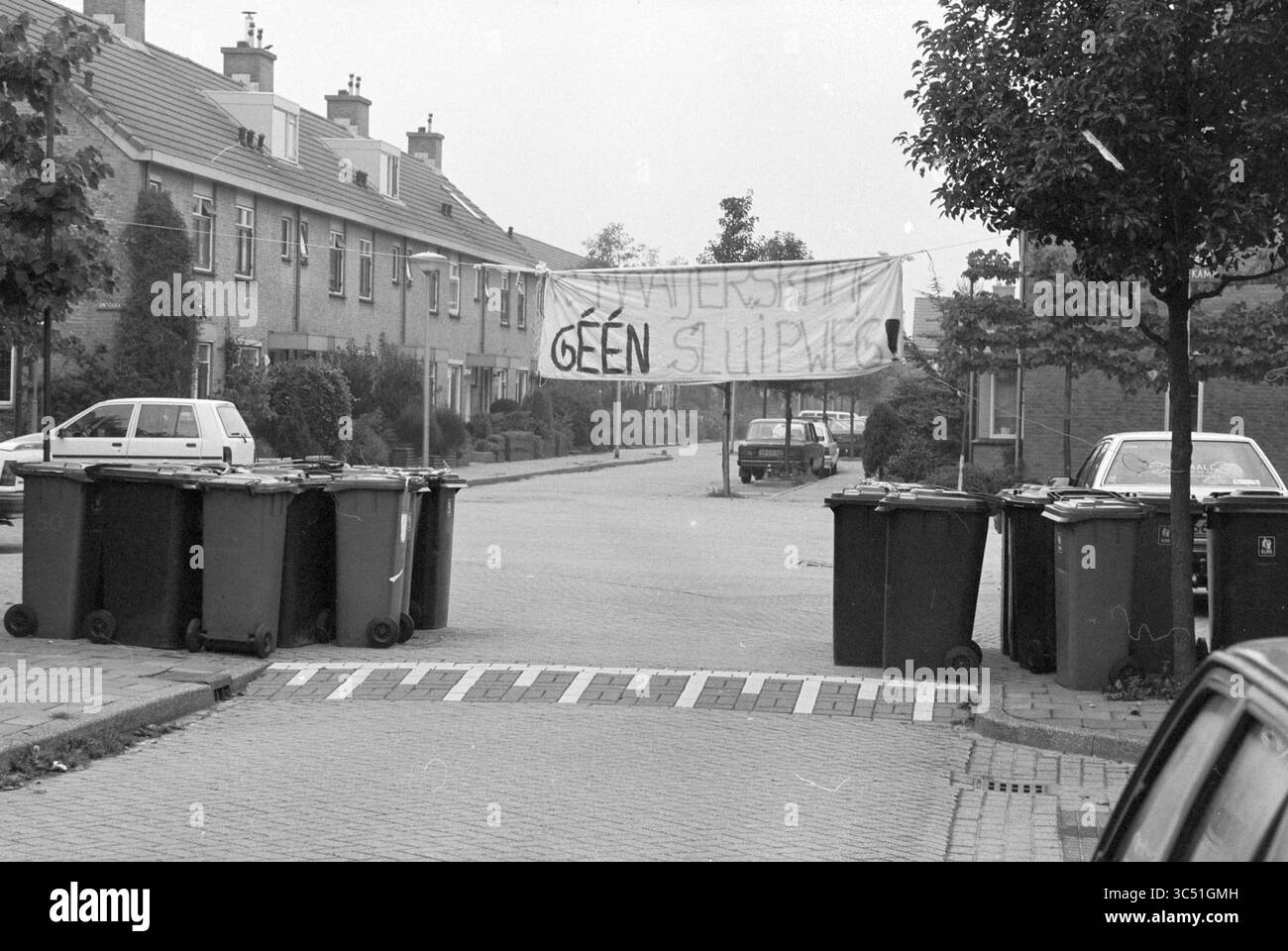 Maßnahmen der Anwohner gegen den Durchgangsverkehr durch Kerkenmaaijerskamp in Velserbroek, Velserbroek, Kerkenmaaijerskamp, 25-09-1994 Whizgle News, Dutch Desk, Niederlande, 1950-2000 Eine Reihe von Mülltonnen säumt die Straße und schafft eine provisorische Barriere. Darüber hängt ein großes Banner zwischen zwei Gebäuden, das kühn eine Botschaft anzeigt. Die Wohngegend wirkt ruhig, mit geparkten Autos und Bäumen an den Seiten. Stockfoto