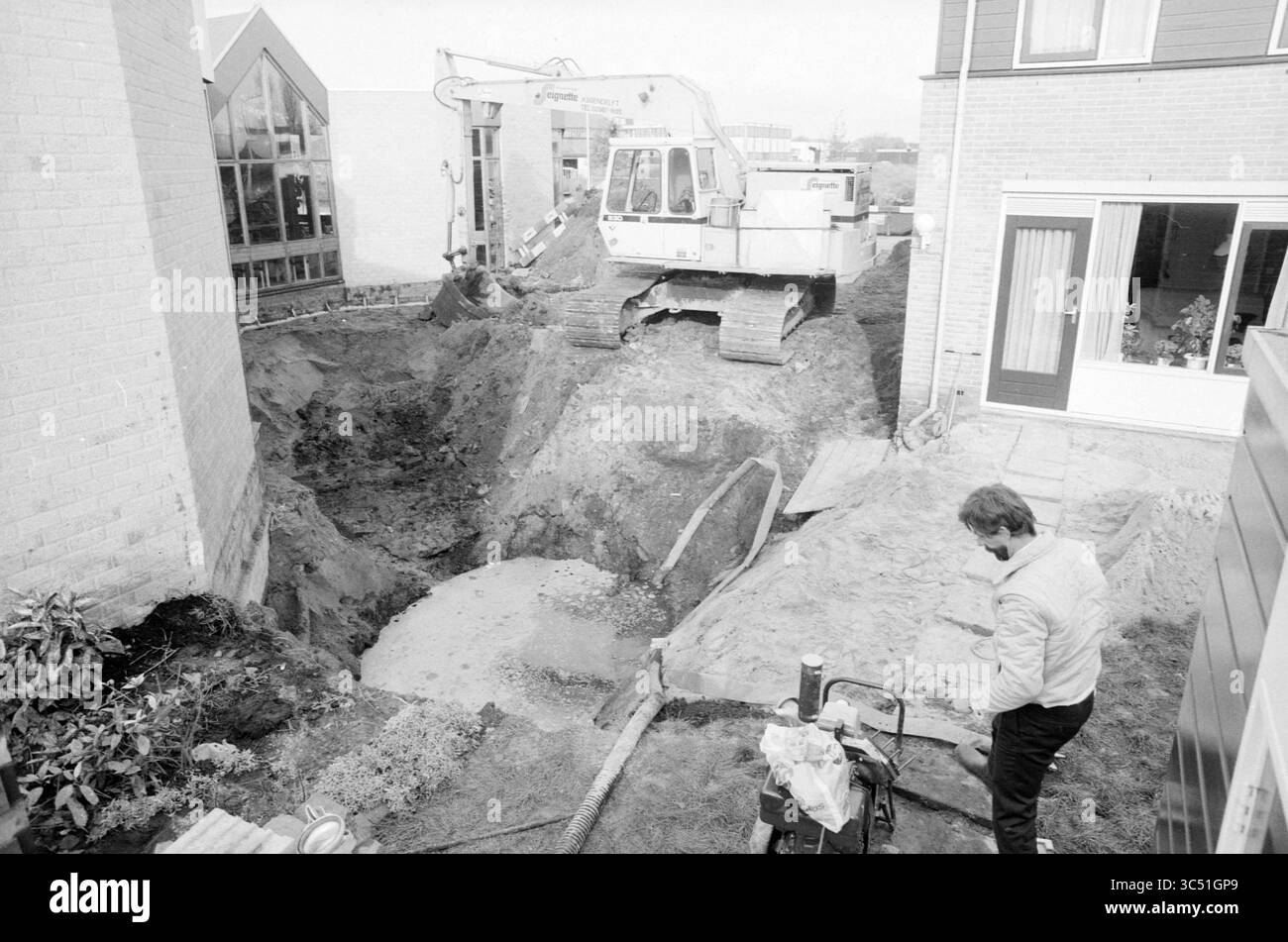 Ausgrabungsarbeiten Hillegom, Work, Hillegom, 04.11-1986 Whizgle News, Dutch Desk, Niederlande, 1950-2000 Auf Einer Baustelle befindet sich ein großer Bagger, der einen tiefen Graben zwischen zwei Gebäuden aushebt, während ein Arbeiter in der Nähe beobachtet. Die Gegend ist von Schmutz und unvollständiger Landschaftsgestaltung umgeben, was die laufenden Renovierungs- oder Bauarbeiten hervorhebt. Stockfoto