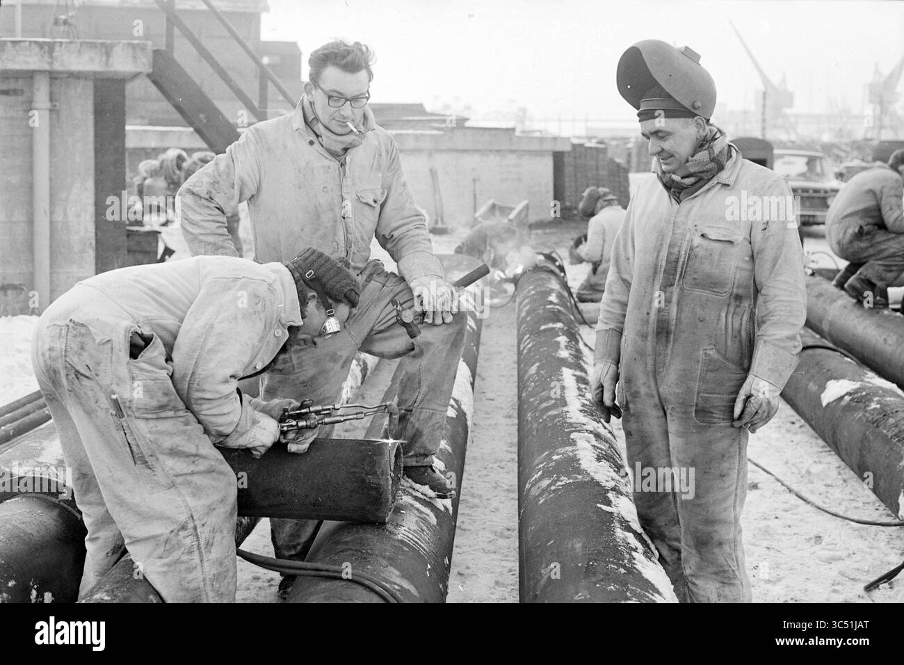 Bau einer Wasserleitung nach Rotterdam in Velsen., Arbeit, 24-01-1963 Whizgle News, Dutch Desk, Niederlande, 1950-2000 Arbeiter in schweren Overalls arbeiten mit Schweißgeräten an großen Rohren am Boden. Ein Mitarbeiter konzentriert sich intensiv auf die Aufgabe, während ein anderer beobachtet, Pfeife im Mund, und ein dritter steht in der Nähe, trägt Schutzkleidung und schaut zu. Im Hintergrund befinden sich industrielle Strukturen und Ausrüstungen. Stockfoto