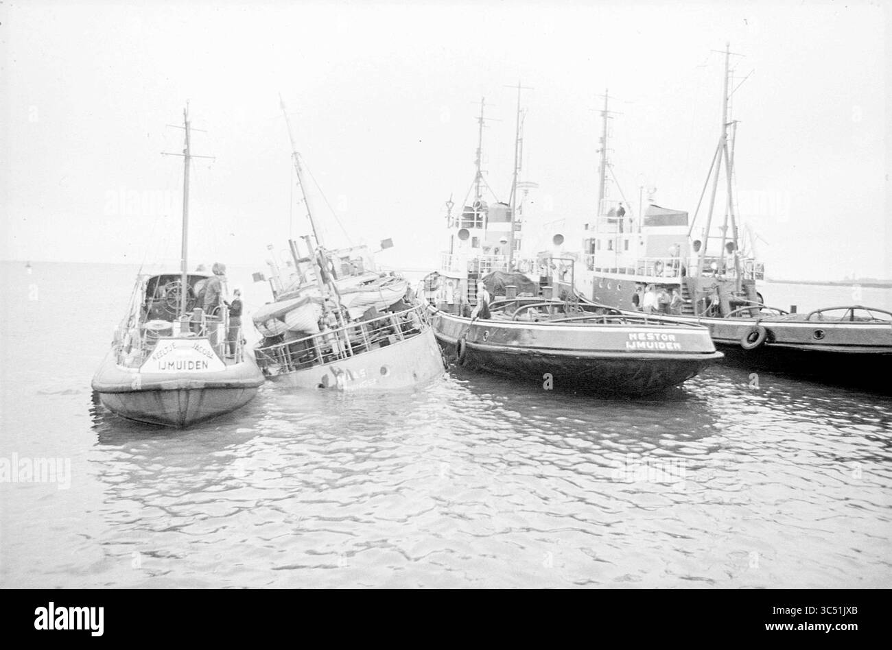 Schleppen in Achterbahn Maas., Wijsmuller, 18-05-1966 Whizgle News, Dutch Desk, Niederlande, 1950-2000 Ein teilweise untergetauchtes Schiff neigt sich prekär zum Wasser, umgeben von mehreren Booten, die in der Nähe verankert sind. Die Besatzungsmitglieder des gelisteten Schiffes scheinen die Situation zu beurteilen, während die ruhigen Gewässer die umliegenden Schiffe und den Himmel widerspiegeln. Stockfoto