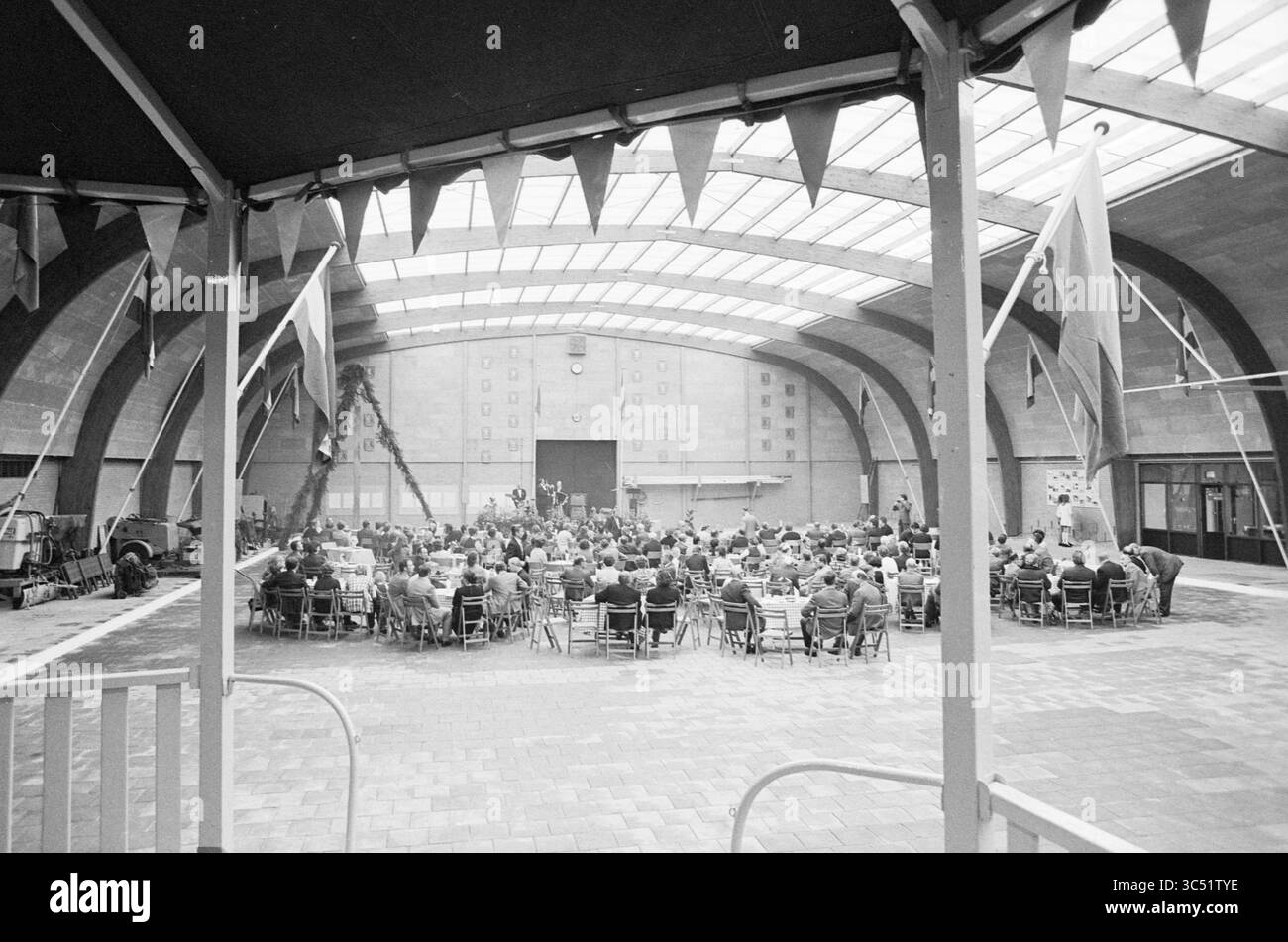 Abteilung für öffentliche Arbeiten, Eröffnungsdepot, Innenraum, Zandvoort, 11-12-1971 Whizgle News, Dutch Desk, Niederlande, 1950-2000 Eine große Versammlung von Personen, die an einem geräumigen, luftigen Ort sitzen und an einer Präsentation oder Veranstaltung teilnehmen. Banner und Fahnen zieren die Decke und verleihen dem Saal einen festlichen Touch, während die Bühne für Lautsprecher oder Aufführungen an der Vorderseite steht. Stockfoto