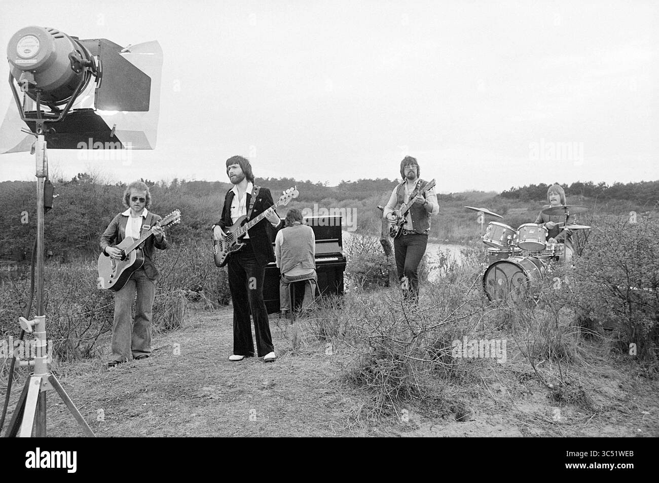 Filmaufnahmen Cats Park Bokkedoorns, Dreharbeiten, Filmaufnahmen, 05-05-1976 Whizgle News, Dutch Desk, Niederlande, 1950-2000 Eine Band tritt im Freien in einer natürlichen Umgebung auf, komplett mit Gitarren, Schlagzeug und Klavier, während sie von einer großen Lichtquelle beleuchtet wird. Die Musiker sind in stilvoller Kleidung gekleidet und präsentieren eine Mischung aus Instrumenten vor einer Kulisse aus Bäumen und freiem Raum. Stockfoto