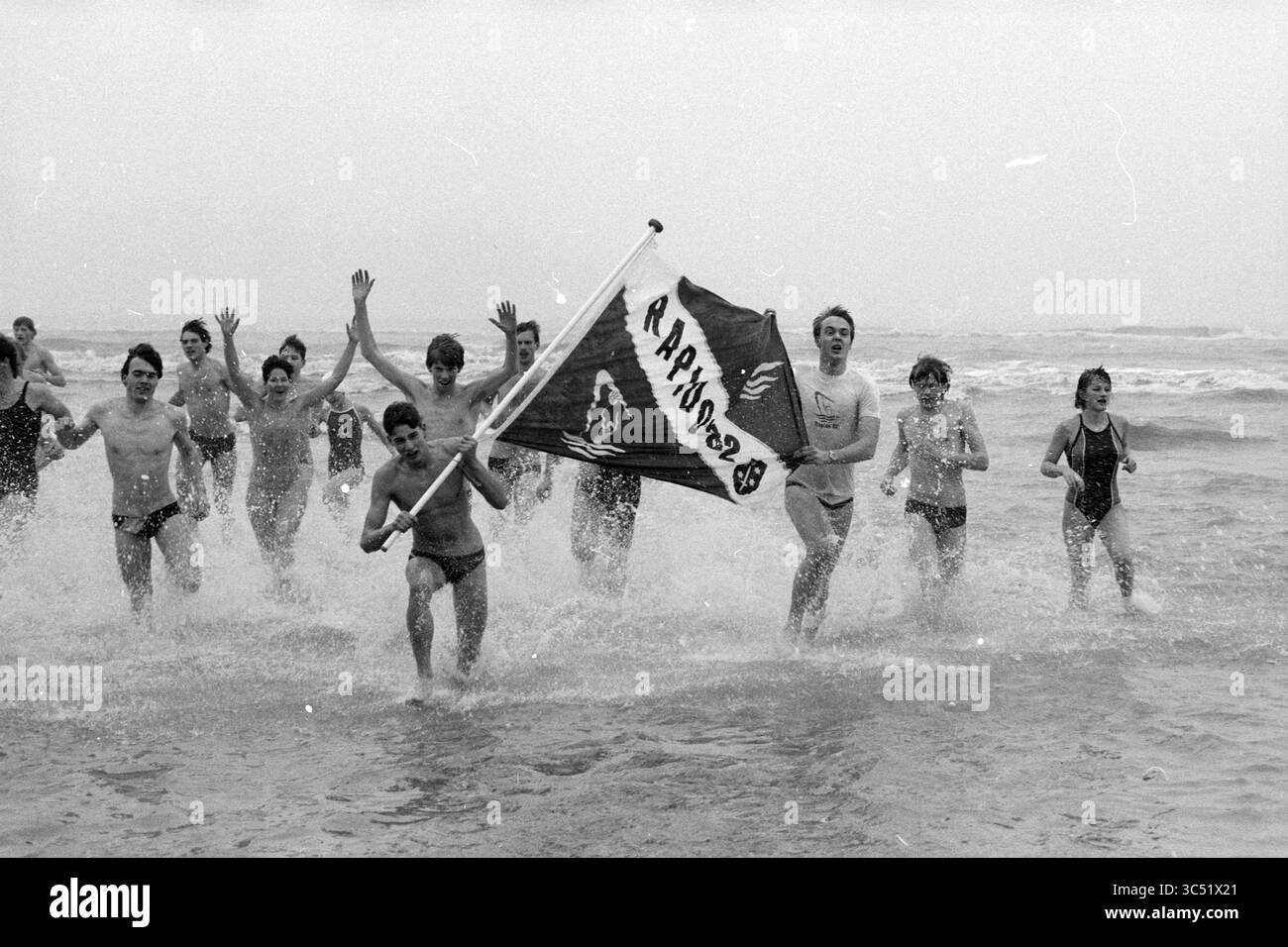Neujahrstauchen Zandvoort, Neujahr, Zandvoort, 01-01-1986 Whizgle News, Dutch Desk, Niederlande, 1950-2000 Eine Gruppe begeisterter Individuen läuft fröhlich durch die Wellen und winkt mit einer Flagge, die mit einem Logo verziert ist. Ihr Lachen und ihre Energie sind spürbar, während sie das erfrischende Meer umarmen. Stockfoto