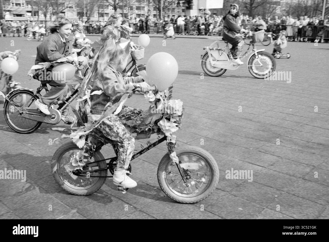 Dekorierte Fahrräder Beverwijk, Queen's Day, Beverwijk, Niederlande, 29-04-1989 Whizgle News, Dutch Desk, Niederlande, 1950-2000 Kinder fahren auf dekorierten Fahrrädern, die mit Ballons und Laub geschmückt sind, während sie an einer lebhaften Parade teilnehmen. Eine fröhliche Menschenmenge beobachtet im Hintergrund und feiert die lebhafte Atmosphäre. Stockfoto