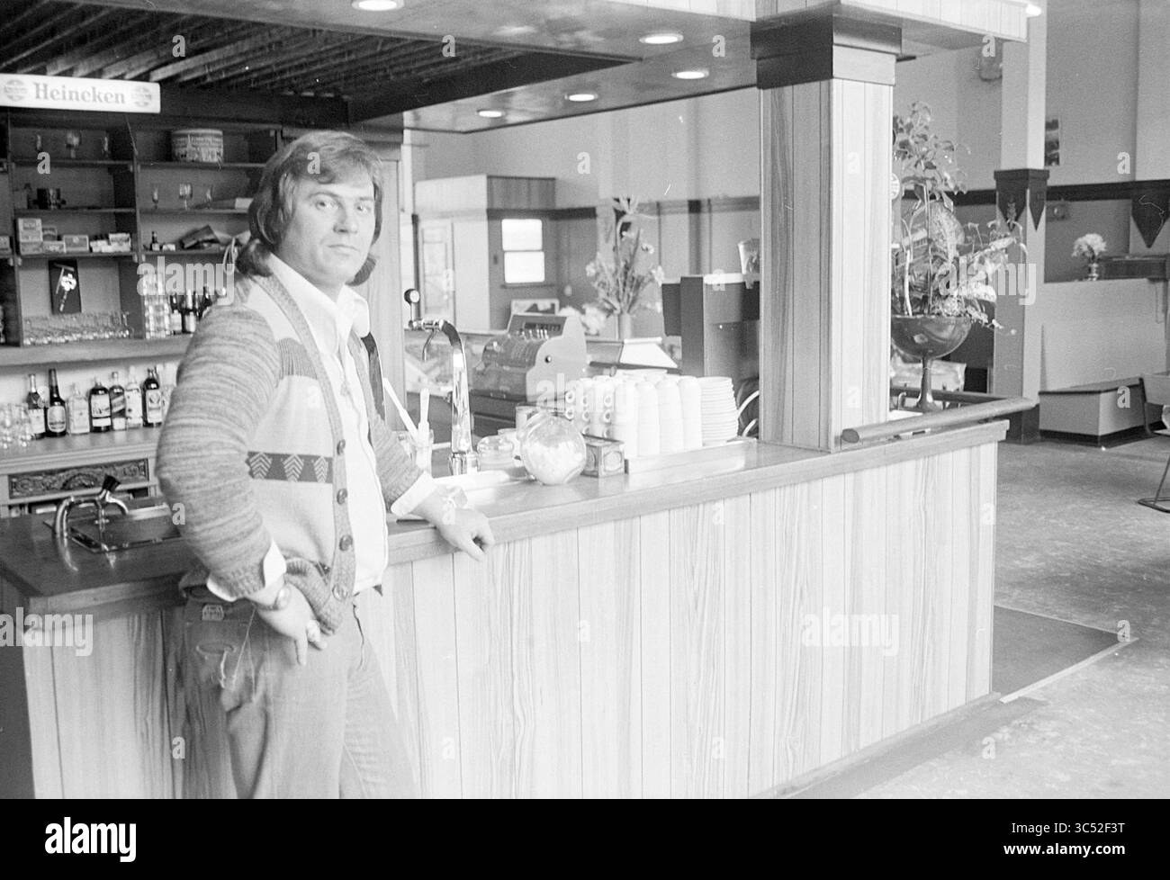 Eigentümer von 'Het Witte Huis', Haven, IJm., Personen, Restaurant, 23-04-1976 Whizgle News, Dutch Desk, Niederlande, 1950-2000 Ein Mann steht selbstbewusst an einer Bar, trägt einen gemusterten Pullover und legere Kleidung. Hinter ihm sind die Regale mit Getränken gefüllt, und die Inneneinrichtung ist schlicht eingerichtet und bietet ein warmes Ambiente. Stockfoto