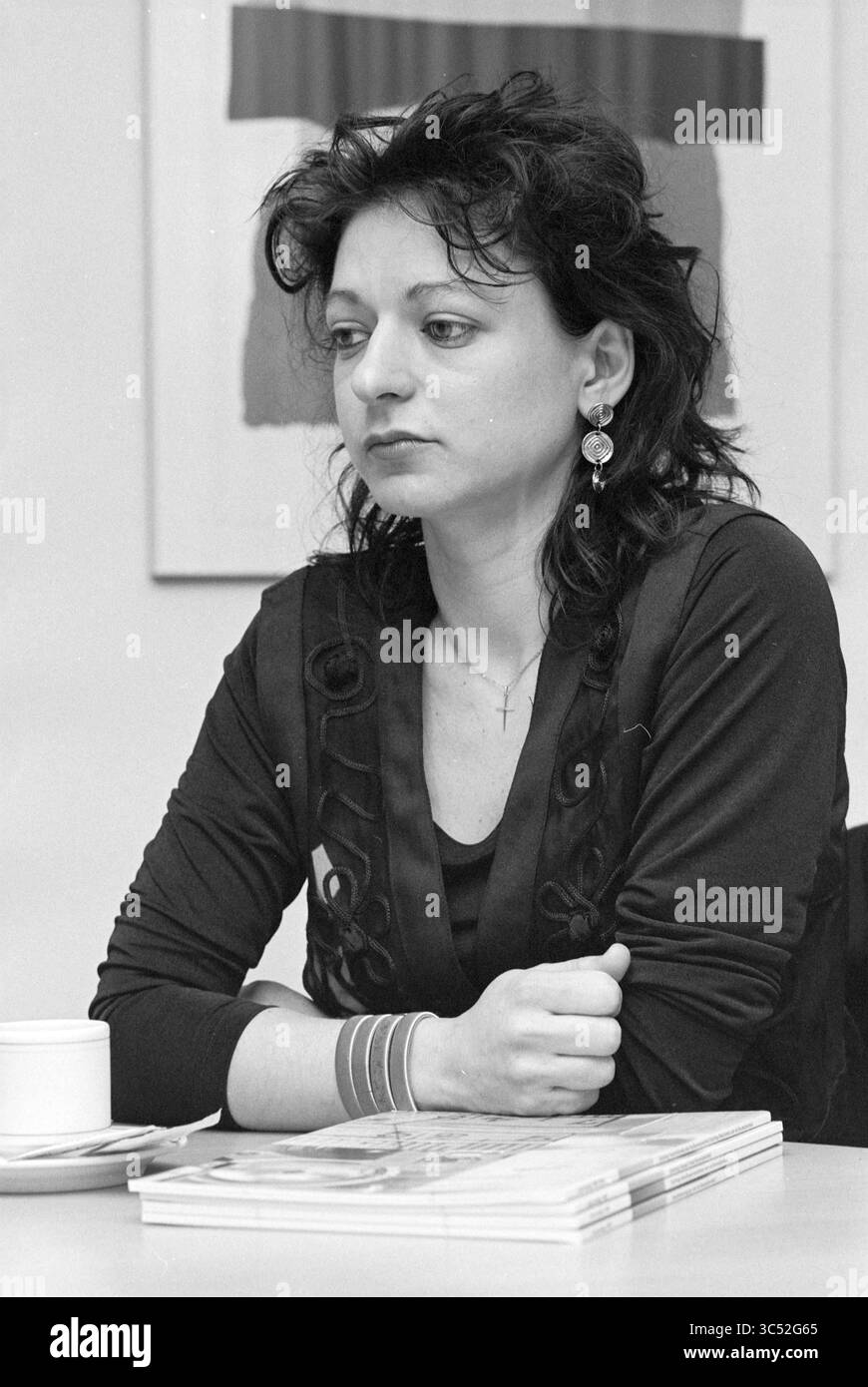 Vier Damen von der Kunsthochschule bei S.F.B., 09-02-1995 Whizgle News, Dutch Desk, Niederlande, 1950-2000 Eine nachdenkliche Frau sitzt an einem Tisch, ihre Arme überkreuzt und ihr Ausdruck besinnlich. Sie trägt unverwechselbare Ohrringe und ein schwarzes Oberteil mit komplizierten Details, umgeben von Magazinen und einer Tasse in der Nähe. Der Hintergrund enthält abstrakte Grafiken, die der Szene eine Ebene Tiefe verleihen. Stockfoto