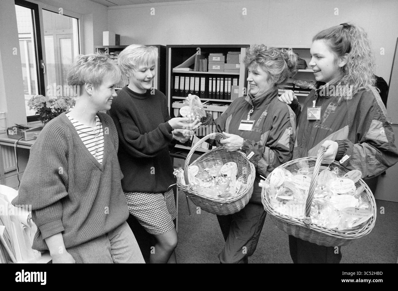 Ostereier verteilen bei der N.A.M., 08-04-1992 Whizgle News, Dutch Desk, Niederlande, 1950-2000 Eine Gruppe von vier Frauen führt ein fröhliches Gespräch, jede hält Körbe voll mit Leckereien. Sie tragen legere Kleidung und zwei Frauen tragen passende Outfits, was auf das Thema Kameradschaft hindeutet. Die Umgebung scheint ein Büro- oder Gemeinschaftsraum zu sein, der einen Moment der gemeinsamen Freude und Verbindung widerspiegelt. Stockfoto