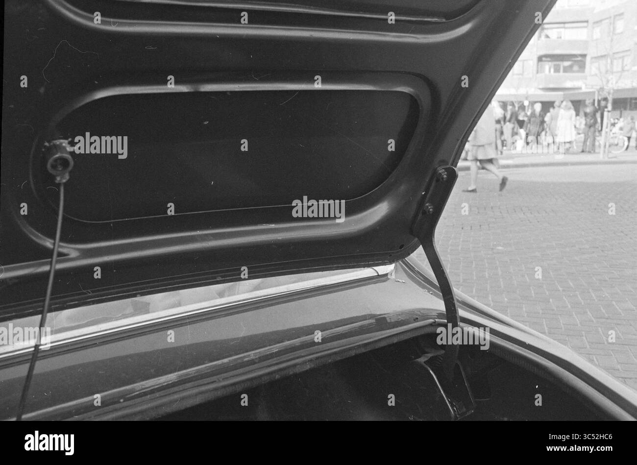 Kofferraum eines Autos Whizgle News, Dutch Desk, Niederlande, 1950-2000 der Kofferraum eines Autos wird geöffnet und enthüllt sein Inneres, während sich im Hintergrund die geschäftige Szene einer Straße voller Fußgänger und Geschäfte entfaltet. Stockfoto