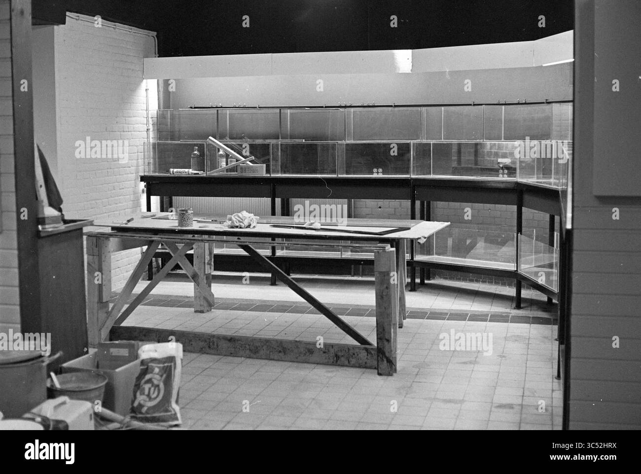 Zwei Männer an einem Arbeitstisch Whizgle News, Dutch Desk, Niederlande, 1950-2000 Ein geräumiger Innenraum zeigt einen unvollendeten Ausstellungsbereich mit eleganten Glasvitrinen, einer Holzwerkbank und verschiedenen Baumaterialien, die über den gefliesten Boden verstreut sind. Die Umwelt deutet auf eine Übergangsphase der Vorbereitung auf ein zukünftiges Schaufenster hin. Stockfoto