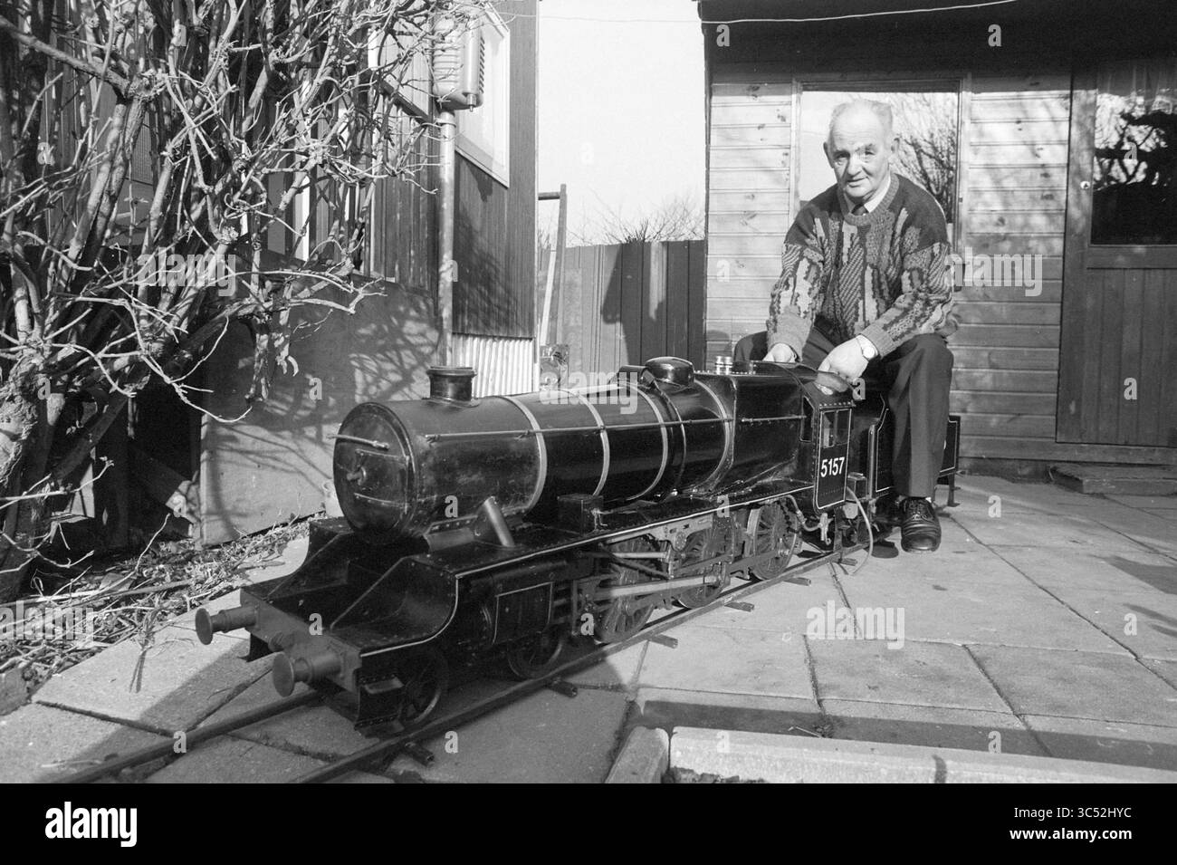 Herr Zwart mit englischer Lokomotive Nieuw-Vennep, Modellbau, Modellbau, Nieuw-Vennep, 08-02-1989 Whizgle News, Dutch Desk, Niederlande, 1950-2000 ein älterer Mann sitzt neben einer Miniatur-Dampflokomotive und zeigt stolz sein kompliziertes Handwerk. Die Umgebung verfügt über einen malerischen Garten mit einem Schuppen im Hintergrund, was eine friedliche Atmosphäre schafft, die den Charme der Modelleisenbahn zum Ausdruck bringt. Stockfoto