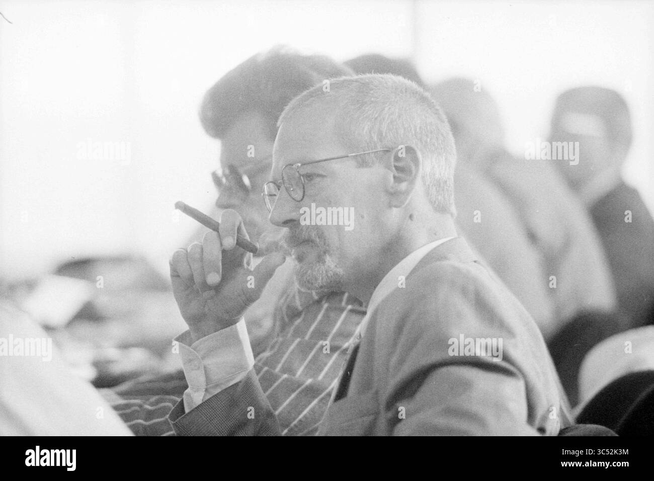 C.O.R. FNV Meeting, 00-00-1990 Whizgle News, Dutch Desk, Niederlande, 1950-2000 Ein nachdenklicher Mann im Anzug, mit Brille und Zigarre, hört während eines Meetings aufmerksam zu, umgeben von anderen, die sich unterhalten. Stockfoto