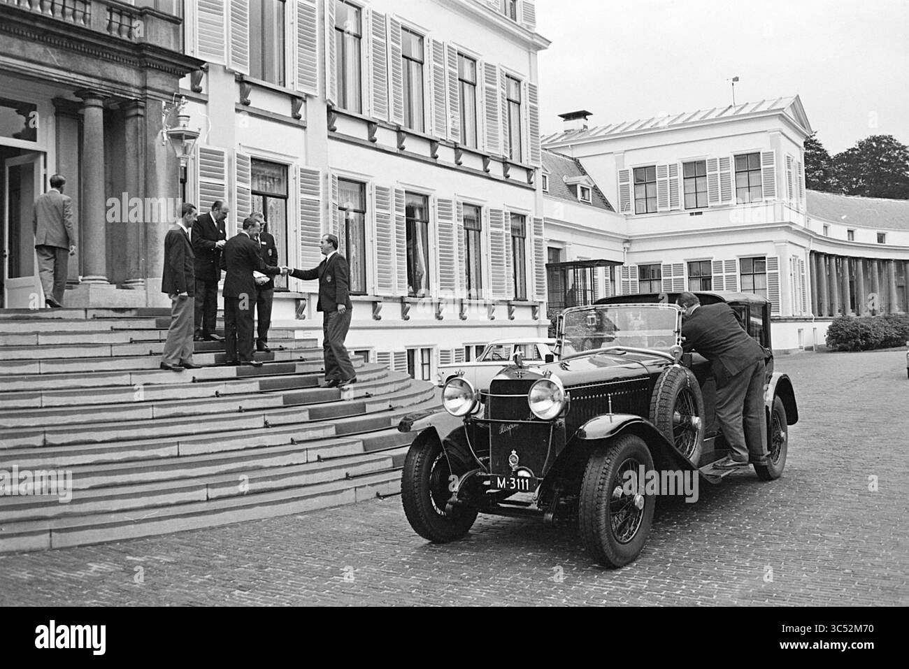 Defile P.A.C. Kundgebung auf Soestdijk für Prinz Bernhard und Lunch Groenendaal, Heemstede, Kon. Besuche und Empfänge, Personen, Beerdigung, 18-09-1965','19-09-1965 Whizgle News, Dutch Desk, Niederlande, 1950-2000 Ein Oldtimer parkt in der Nähe eines großen Gebäudes, während sich einige Leute auf den Stufen unterhalten. Die Szene fängt einen Moment der Interaktion ein, in dem eine Person scheinbar andere begrüßt. Die Architektur verfügt über große Fenster und Fensterläden, die zur eleganten Atmosphäre der Umgebung beitragen. Stockfoto