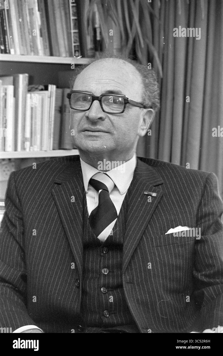 Porträt Mr. Van der Waal Heemstede - Planungsdienst, Porträts, Porträts, 15-04-1975 Whizgle News, Dutch Desk, Niederlande, 1950-2000 Ein Mann in formeller Kleidung, Brille und vor einem Bücherregal sitzend, strahlt ein nachdenkliches Verhalten aus. Sein Nadelstreifenanzug und die sorgfältig angeordnete Krawatte lassen eine professionelle Atmosphäre vermuten, während sein leichtes Lächeln auf eine einladende Persönlichkeit hinweist. Stockfoto