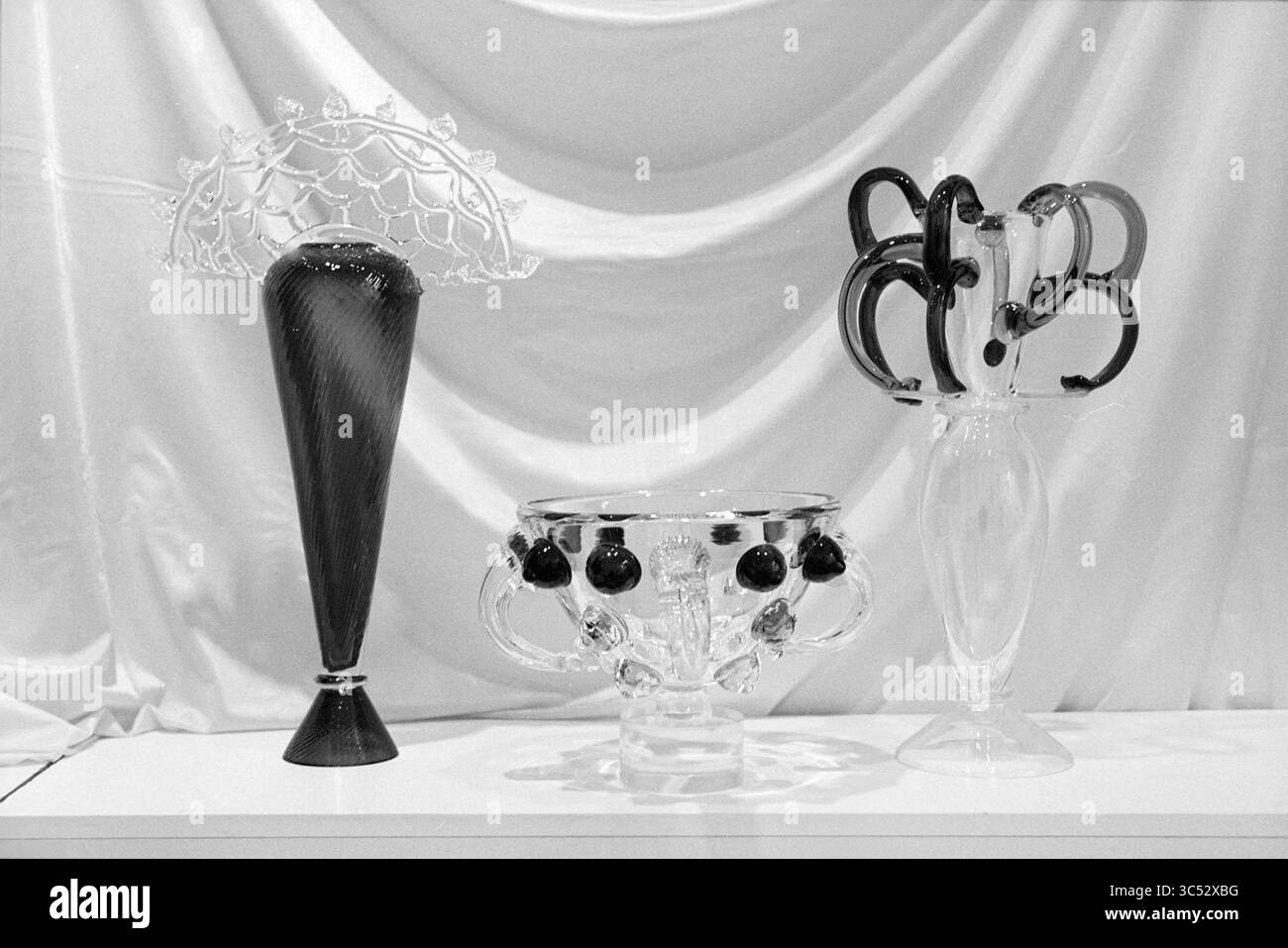 Glasobjekte, Sibek, 09-03-1995 Whizgle News, Dutch Desk, Niederlande, 1950-2000 drei kunstvoll verzierte Glasvasen zeigen eine Mischung aus komplexen Designs und Formen. Die linke Vase hat eine hohe, schlanke Form mit zarten Details, während das mittlere Teil einen abgerundeten Körper mit dunklen kugelförmigen Akzenten aufweist. Die rechte Vase zeigt einen eher skulpturalen Stil, gekennzeichnet durch gewellte Griffe, die eine skurrile Note verleihen. Alle Vasen sind auf einem weich drapierten Hintergrund angeordnet und betonen ihre Eleganz. Stockfoto