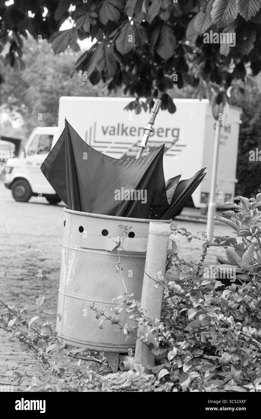 Regenschirm im Mülleimer, Verschiedenes, 02-08-1989 Whizgle News, Dutch Desk, Niederlande, 1950-2000 ein verlassener schwarzer Regenschirm liegt kopfüber in einem Abfalleimer aus Metall, umgeben von Grün, während ein Lieferwagen im Hintergrund parkt. Stockfoto