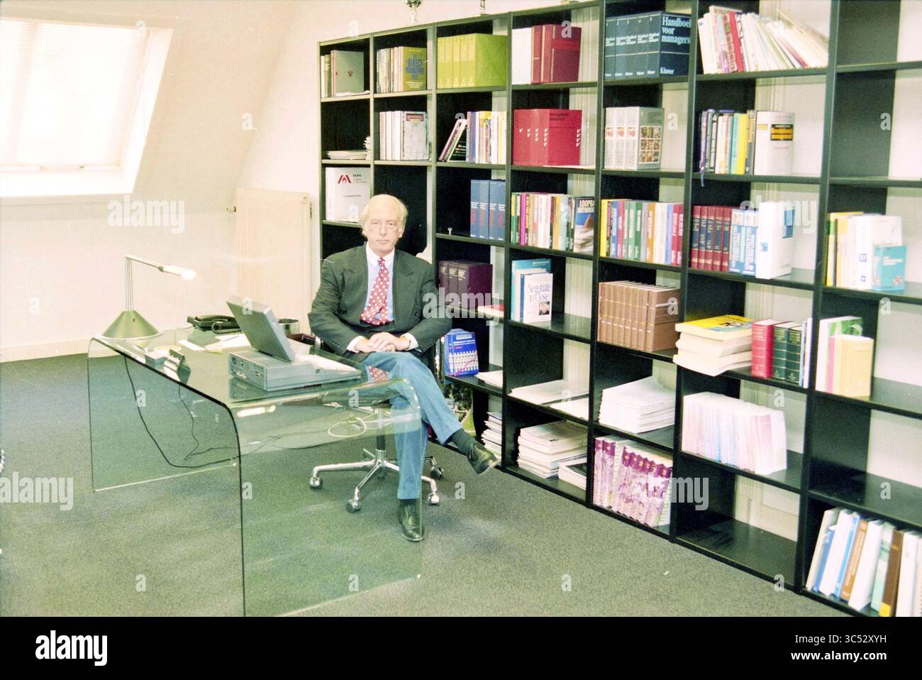 Jean Smith, 30-01-1996 Whizgle News, Dutch Desk, Niederlande, 1950-2000 Eine angesehene Person sitzt an einem eleganten Glasschreibtisch in einem modernen Büro, umgeben von ordentlich organisierten Bücherregalen, gefüllt mit verschiedenen Büchern und Ordnern, was eine wissenschaftliche Atmosphäre schafft. Stockfoto