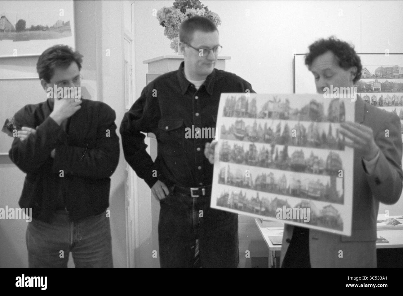 Enthüllung von Postkarten, 16-12-1994 Whizgle News, Dutch Desk, Niederlande, 1950-2000 drei Personen sind an einer Diskussion beteiligt, wobei einer von ihnen einen großen Druck mit einer Reihe detaillierter Illustrationen von Gebäuden in der Hand hält. Die anderen beiden beobachten aufmerksam, mit einer Hand am Kinn, tief in Gedanken. Die Umgebung scheint eine Ausstellung oder Galerie zu sein, die eine Atmosphäre künstlerischer Auseinandersetzung und Kritik widerspiegelt. Stockfoto