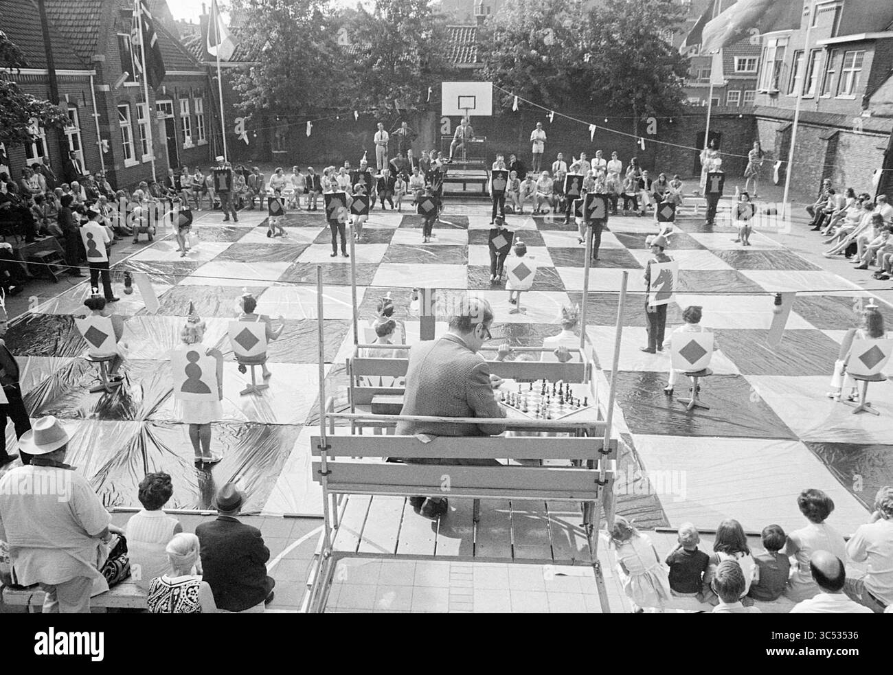 Living Chess Game Bomans - Euwe, Chess, 29-08-1970 Whizgle News, Dutch Desk, Niederlande, 1950-2000 in einem Innenhof wird Ein großes Freischachbrett aufgestellt, in dem die als Schachfiguren gekleideten Teilnehmer ihre Positionen einnehmen. Zuschauer umgeben das Gebiet und beobachten das einzigartige Spiel. Ein Mann, der auf einer hohen Plattform sitzt, überwacht das Spiel, tief in Gedanken, während farbenfrohe Banner der Veranstaltung eine festliche Atmosphäre verleihen. Stockfoto