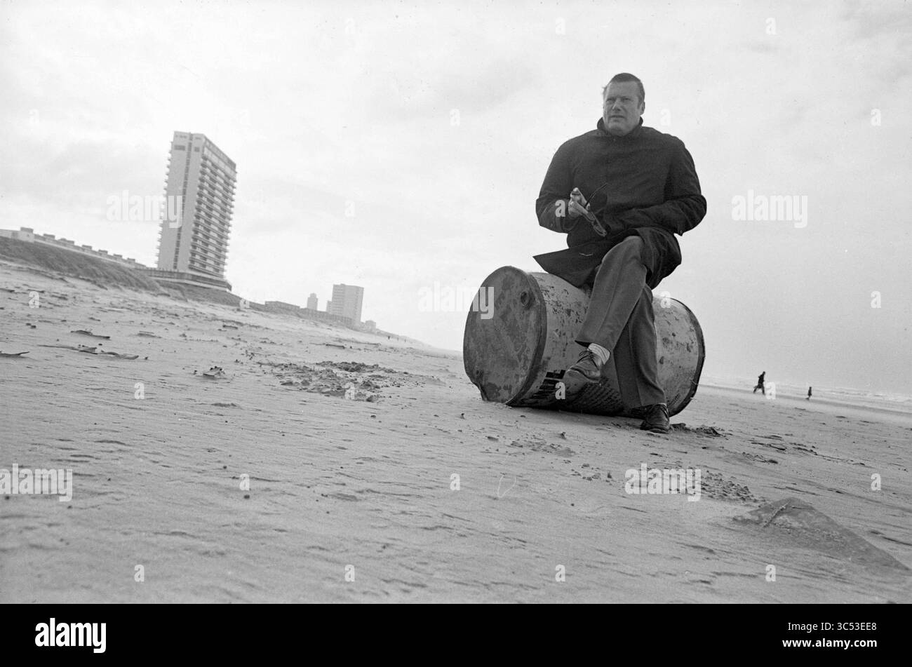 Mr. Ruhling am Strand, Boxen, Personen, 22-10-1970 Whizgle News, Dutch Desk, Niederlande, 1950-2000 Ein Mann sitzt auf einem großen Fass an einem windgepeitschten Strand und blickt nachdenklich in die Ferne. Im Hintergrund erhebt sich ein modernes Gebäude vor einem bewölkten Himmel, während einige Figuren am Ufer entlang spazieren. Die Szene fängt eine Mischung aus Einsamkeit und urbanem Leben an der Küste ein. Stockfoto