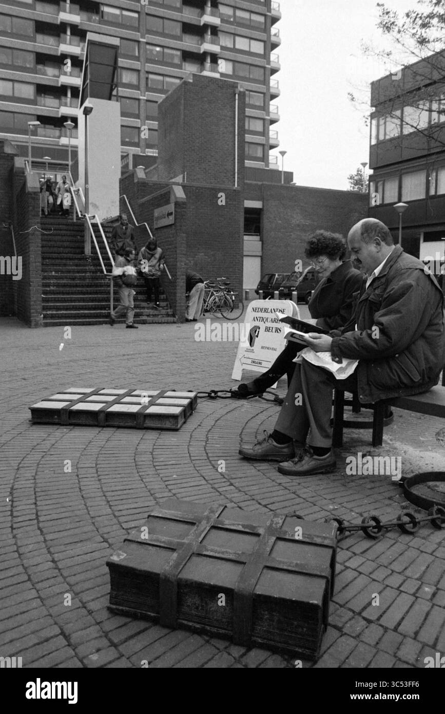 Große Bücher, Stationsplein, Haarlem, Stationsplein, Niederlande, 31-10-1991, Whizgle News, Dutch Desk, Niederlande, 1950-2000 zwei Personen sitzen auf einer Bank und erscheinen tief in Gedanken, während große, angekettete Kisten auf dem Boden in der Nähe liegen. Im Hintergrund führt eine Treppe zu einem mehrstöckigen Gebäude, und in der Ferne ist eine kleine Menschenmenge zu sehen, die der Szene ein Gefühl von geschäftiger Aktivität verleiht. Ein Schild in der Nähe weist auf eine Gemeindeveranstaltung oder ein Treffen hin. Stockfoto