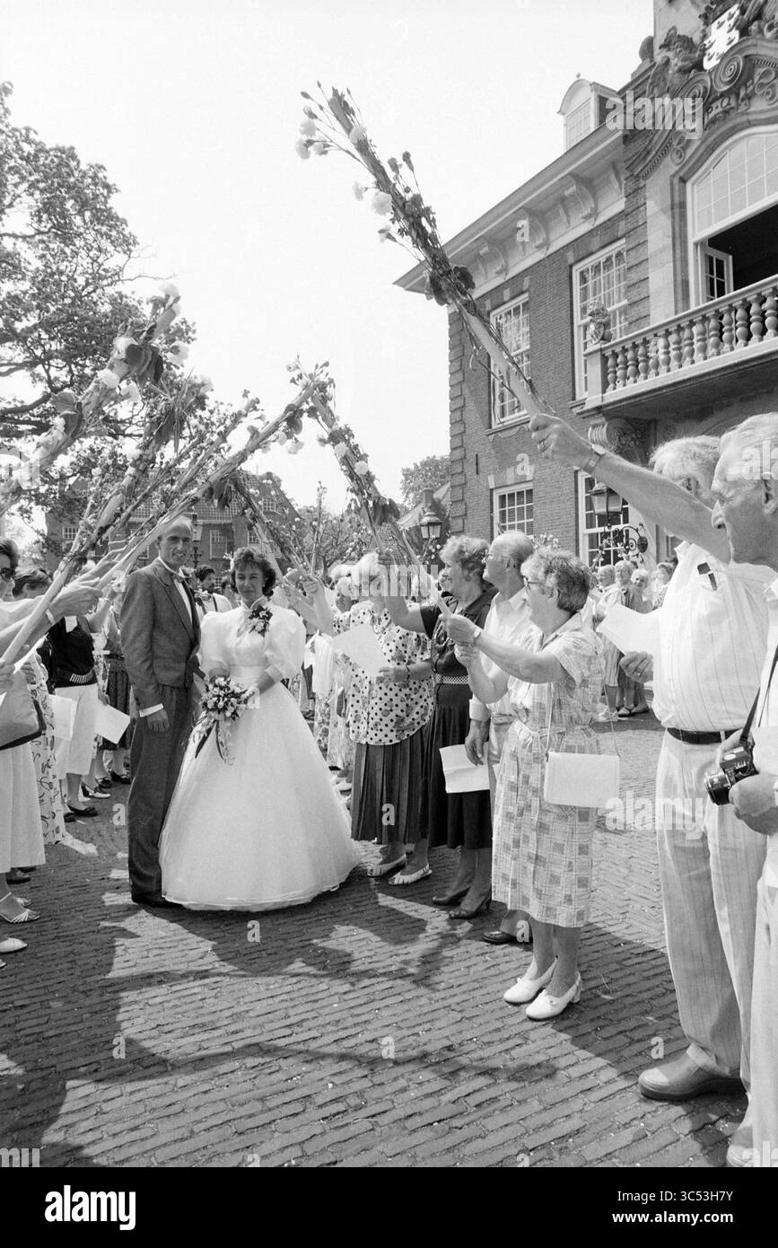 Heirat Rettungsschwimmer Hopman Heemstede, Heirat, Heemstede, Niederlande, 19-05-1989 Whizgle News, Dutch Desk, Niederlande, 1950-2000 Eine Braut und ein Bräutigam stehen zusammen, umgeben von einer fröhlichen Menschenmenge, die Blumensträuße und Blumen hält, um ihren besonderen Tag zu feiern. Die Gäste, die in verschiedenen Outfits gekleidet sind, lächeln, während sie mit den Blumenarrangements einen festlichen Bogen kreieren. Stockfoto