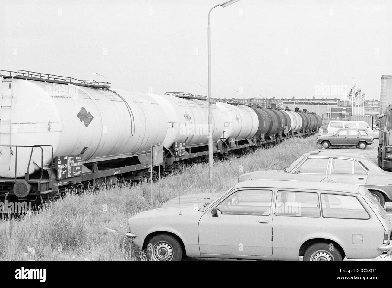 Zugwagen mit Gas, stationär in Kromhoutstraat, IJmuiden, IJmuiden, Kromhoutstraat, Niederlande, 18-08-1981 Whizgle News, Dutch Desk, Niederlande, 1950-2000 Eine Reihe von Tankwagen, die neben vielen Fahrzeugen geparkt sind, wobei Gras und Unkraut zwischen den Bahngleisen und geparkten Autos wachsen, was auf eine industrielle Umgebung hindeutet. Stockfoto