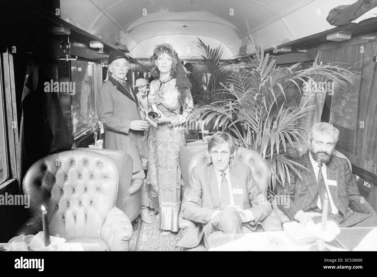 Ankunft Orient Express in Haarlem, Trains, Haarlem, Niederlande, 19-03-1986 Whizgle News, Dutch Desk, Niederlande, 1950-2000 ein elegantes Treffen in einem üppigen Eisenbahnwagen mit vier Personen: ein Mann in einem Anzug mit einer kleinen Trophäe, eine Frau in einem detailverliebten Kleid und zwei weitere Männer, die sich in üppigem Grün und kunstvollen Möbeln unterhalten. Stockfoto