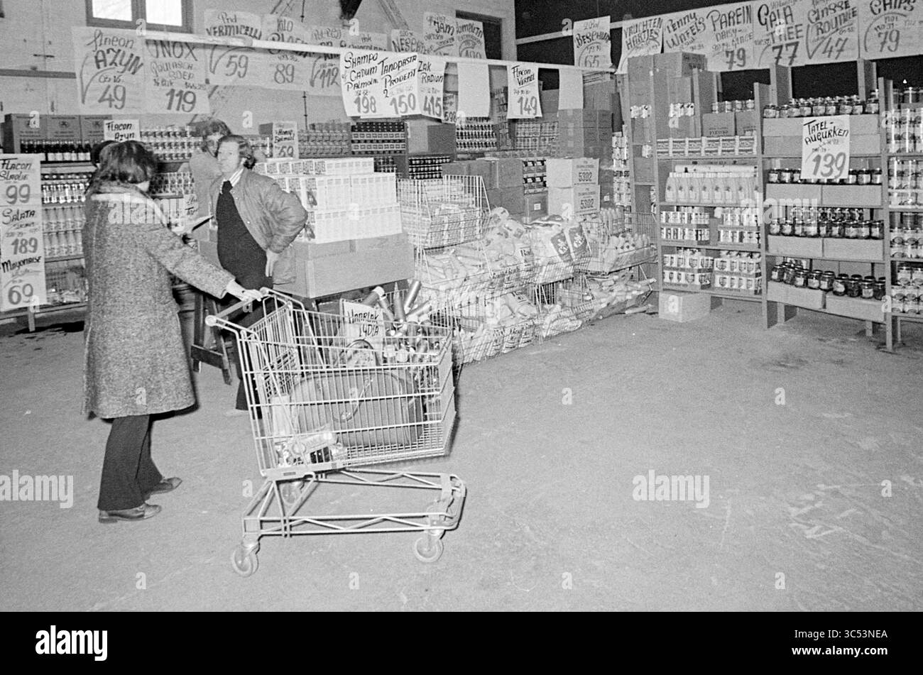 Cash & Carry Shopping Whizgle News, Dutch Desk, Niederlande, 1950-2000 Ein belebtes Lebensmittelladen mit Regalen, die mit verschiedenen Dosenwaren und verpackten Artikeln gestapelt sind. Käufer durchstöbern die Gänge, während die Preise auf Schildern im gesamten Raum deutlich sichtbar angezeigt werden, was das Gefühl vermittelt, auf einem lokalen Markt in der Gemeinschaft einzukaufen. Stockfoto