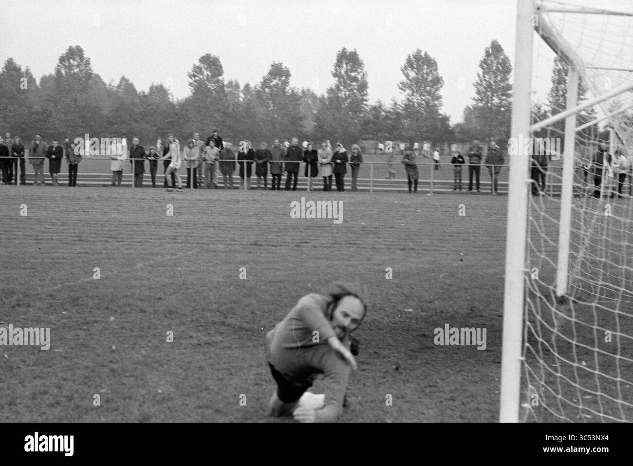 Football Match Whizgle News, Dutch Desk, Niederlande, 1950-2000 Ein Torhüter taucht Ein, um sich zu retten, während eine Menschenmenge hinter einem Zaun, umgeben von Bäumen im Hintergrund, aufmerksam zusieht. Stockfoto