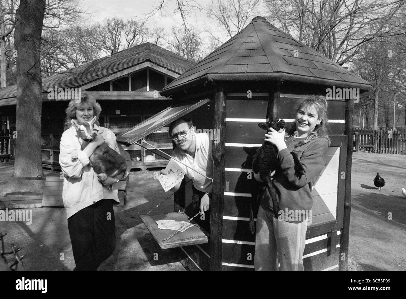 Füttern Sie Kinder Groenendaal, Animals, Groenendaal, Heemstede, Niederlande, 29-03-1989 Whizgle News, Dutch Desk, Niederlande, 1950-2000 Eine Frau hält ein kleines Kaninchen, während eine andere Frau und ein Mann spielerisch in einer malerischen Holzstruktur posieren, die jeweils eine Katze wiegt. Um sie herum sind Bäume und ein Blick auf eine Parklandschaft. Stockfoto