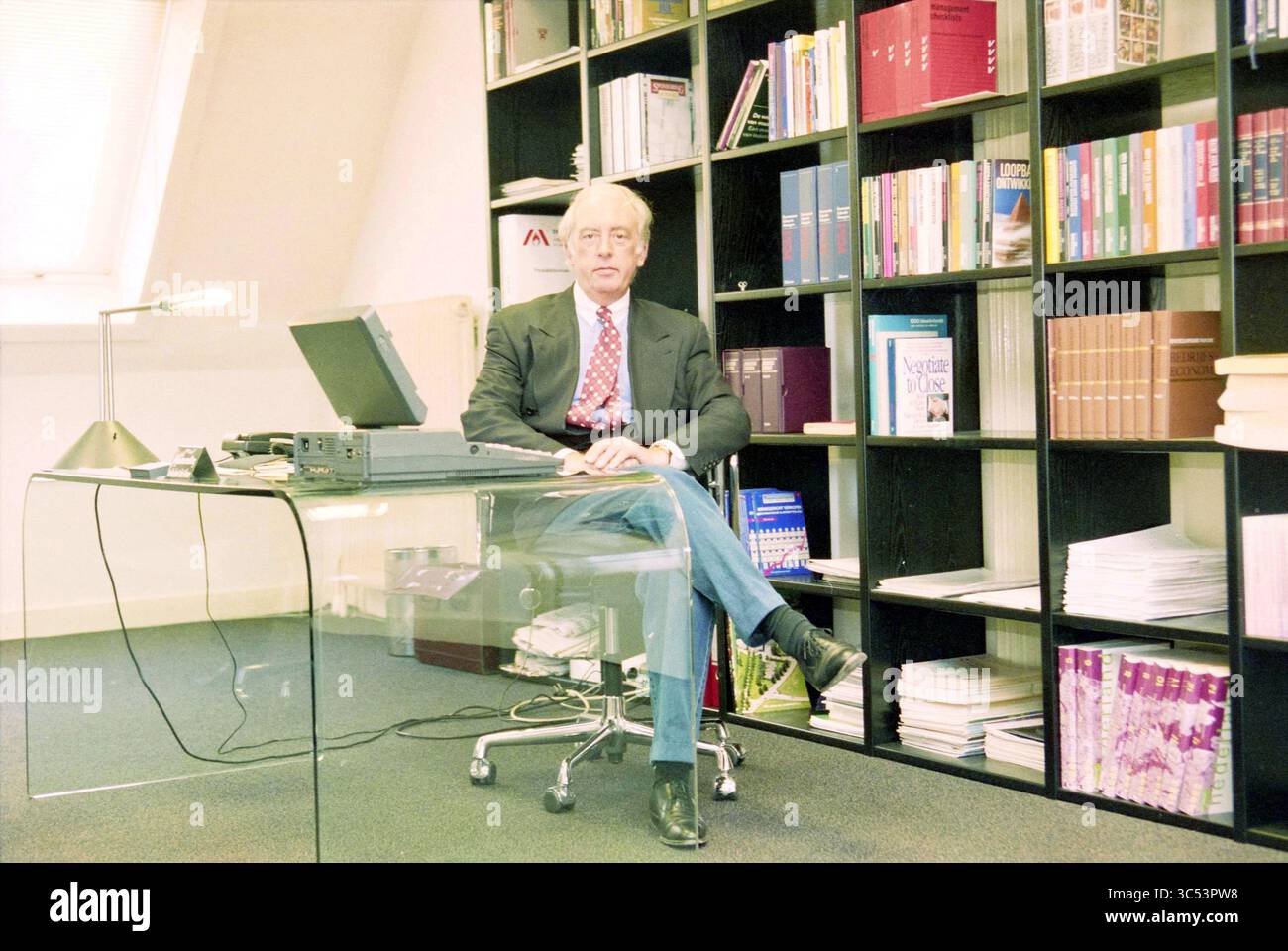 Jean Smith, 30-01-1996 Whizgle News, Dutch Desk, Niederlande, 1950-2000 Ein Mann im Anzug sitzt an einem modernen, klaren Schreibtisch, der vor einem Bücherregal mit verschiedenen Büchern und Organisationsmaterialien steht. Das Büro verfügt über ein professionelles Ambiente mit einer Lampe und einem kleinen elektronischen Gerät, das die Funktionalität des Arbeitsplatzes erhöht. Stockfoto