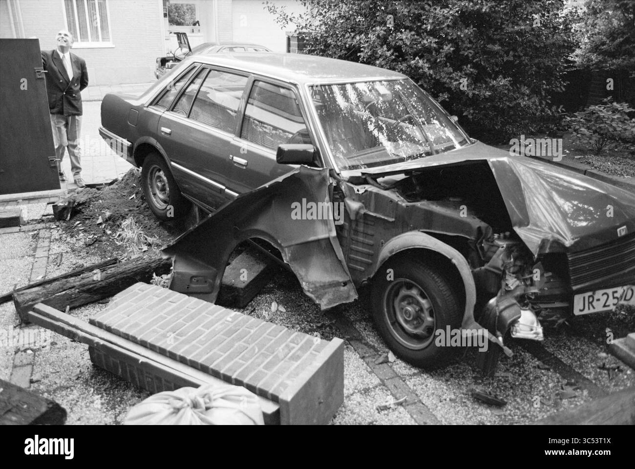 Auto in Dr. Karsten's Garten, 01-10-1994 Whizgle News, Dutch Desk, Niederlande, 1950-2000 Ein beschädigtes Auto liegt teilweise von einer Auffahrt entfernt, mit einem gebogenen vorderen Ende und einem umgefallenen Baumstamm in der Nähe. Im Hintergrund steht eine Person, die die Szene mit einem leicht amüsanten Ausdruck betrachtet. In der Umgebung gibt es verstreute Ziegelsteine und Grünflächen. Stockfoto