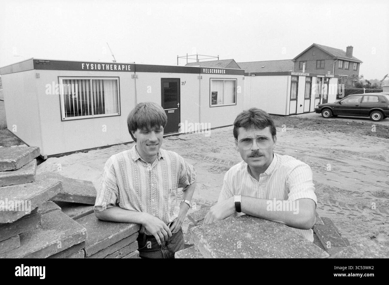 2 Therapeuten Velserbroek, Personen Krankenhaus Krankenpflege, Velserbroek, 01-09-1988 Whizgle News, Dutch Desk, Niederlande, 1950-2000 zwei Männer stehen vor einem modularen Gebäude mit der Bezeichnung „Fysiotherapie“ und „Verpleging“, im Hintergrund mit einer Baustelle und einem Haus. Ein Mann hat kurze Haare und trägt ein gestreiftes Hemd, während der andere eine Brille trägt und einen Schnurrbart trägt. Beide scheinen in Gesprächen zu sein. Gepflasterte Steine sind in der Nähe stapelbar, was auf laufende Arbeiten in der Gegend hindeutet. Stockfoto