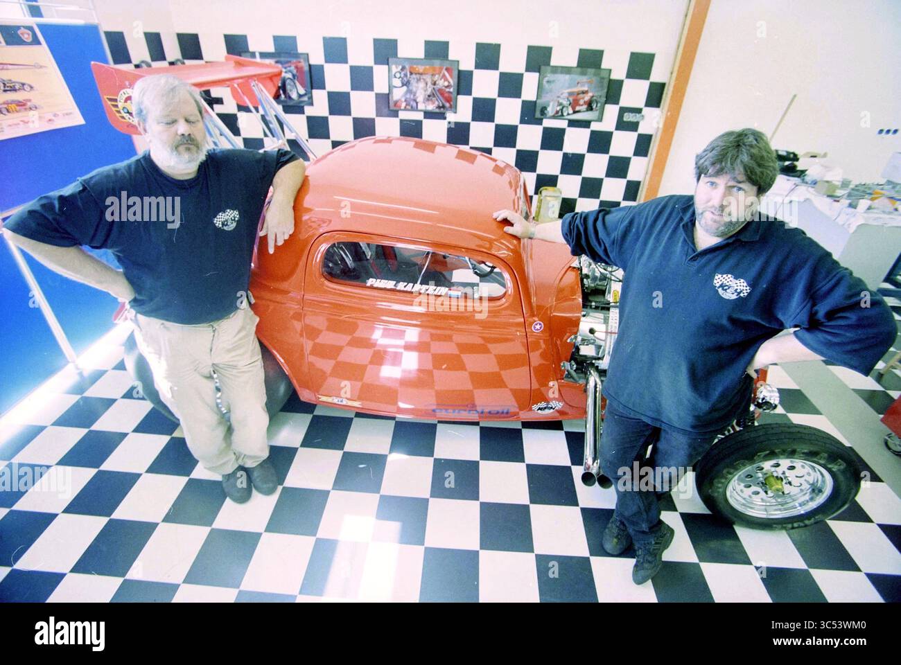 K&H Racing Team IJmuiden, IJmuiden, Niederlande, 17-04-2001 Whizgle News, Dutch Desk, Niederlande, 1950-2000 zwei Männer stehen neben einem Oldtimer-Rennwagen und strahlen ein entspanntes Selbstvertrauen aus. Das Fahrzeug verfügt über ein glänzendes rotes Äußere mit markanten Rennaufklebern, während die Kulisse ein kariertes Muster in Schwarz-weiß zeigt. Der Raum ist voller Leidenschaft für die Automobilbranche, die durch die Werkzeuge und Erinnerungsstücke um sie herum hervorgehoben wird. Stockfoto