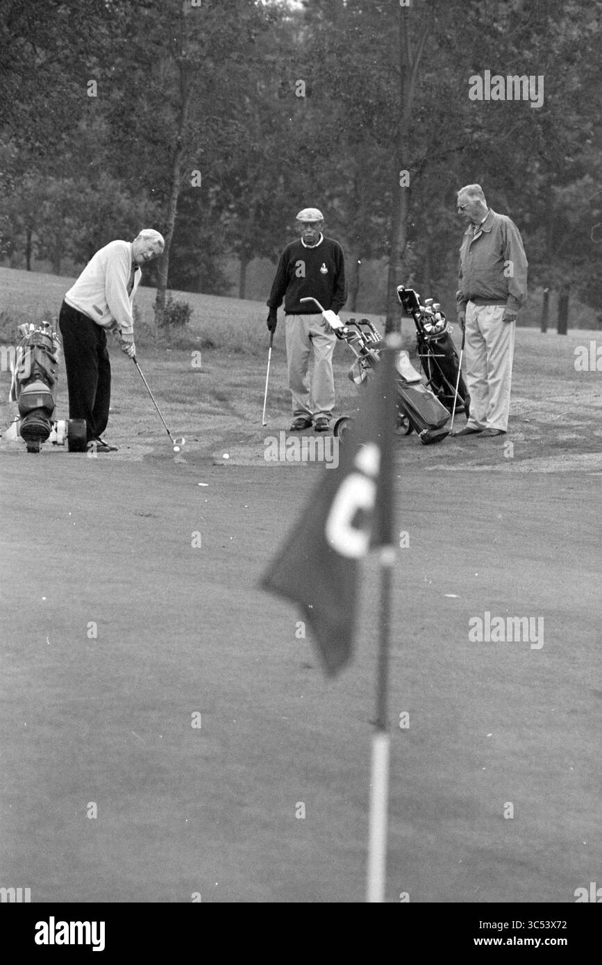 Erfahrene ältere Menschen: Golf, Golfplatz, 11-09-1995 Whizgle News, Dutch Desk, Niederlande, 1950-2000 drei Golfspieler spielen auf dem Green ein Freundschaftsspiel, wobei einer sich auf den Putt vorbereitet, während die anderen beobachten. Golftaschen befinden sich in der Nähe, und eine Flagge markiert das Loch im Vordergrund, was die ruhige Atmosphäre des Platzes unterstreicht. Stockfoto