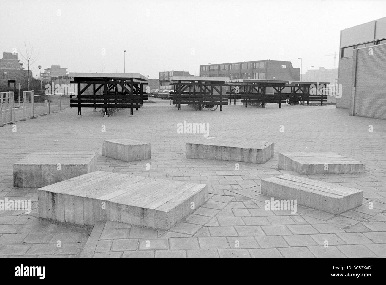 Kunstwerk + Außenansicht Christina MAVO, Schulen, 11-01-1980 Whizgle News, Dutch Desk, Niederlande, 1950-2000 Ein weitläufiger Stadtbereich verfügt über mehrere eckige Betonplattformen, die kreisförmig angeordnet sind und von Holzkonstruktionen umgeben sind, die als Unterschlupf oder Sitzgelegenheit dienen. Die Kulisse zeigt eine Mischung von Gebäuden, die die industrielle Atmosphäre der Umgebung noch verstärken. Stockfoto