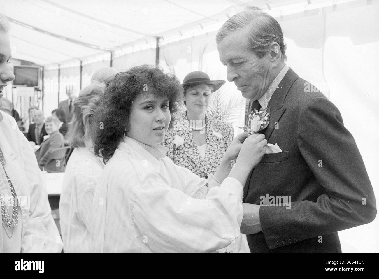 Eröffnung der Dampfpumpenstation auf halbem Weg durch Prince Claus, Eröffnung, Eröffnung, Inbetriebnahme, Dampfpumpenstation und Dampfpumpenstation, Halfweg, 10-06-1987 Whizgle News, Dutch Desk, Niederlande, 1950-2000 Eine junge Frau steckt einem elegant gekleideten Mann eine Blume zart an das Revers, der scheinbar in Gesprächen verwickelt ist. Hinter ihnen versammelt sich eine Gruppe von Menschen, darunter eine Frau mit Hut und andere in formeller Kleidung, um eine festliche Atmosphäre zu schaffen. Stockfoto