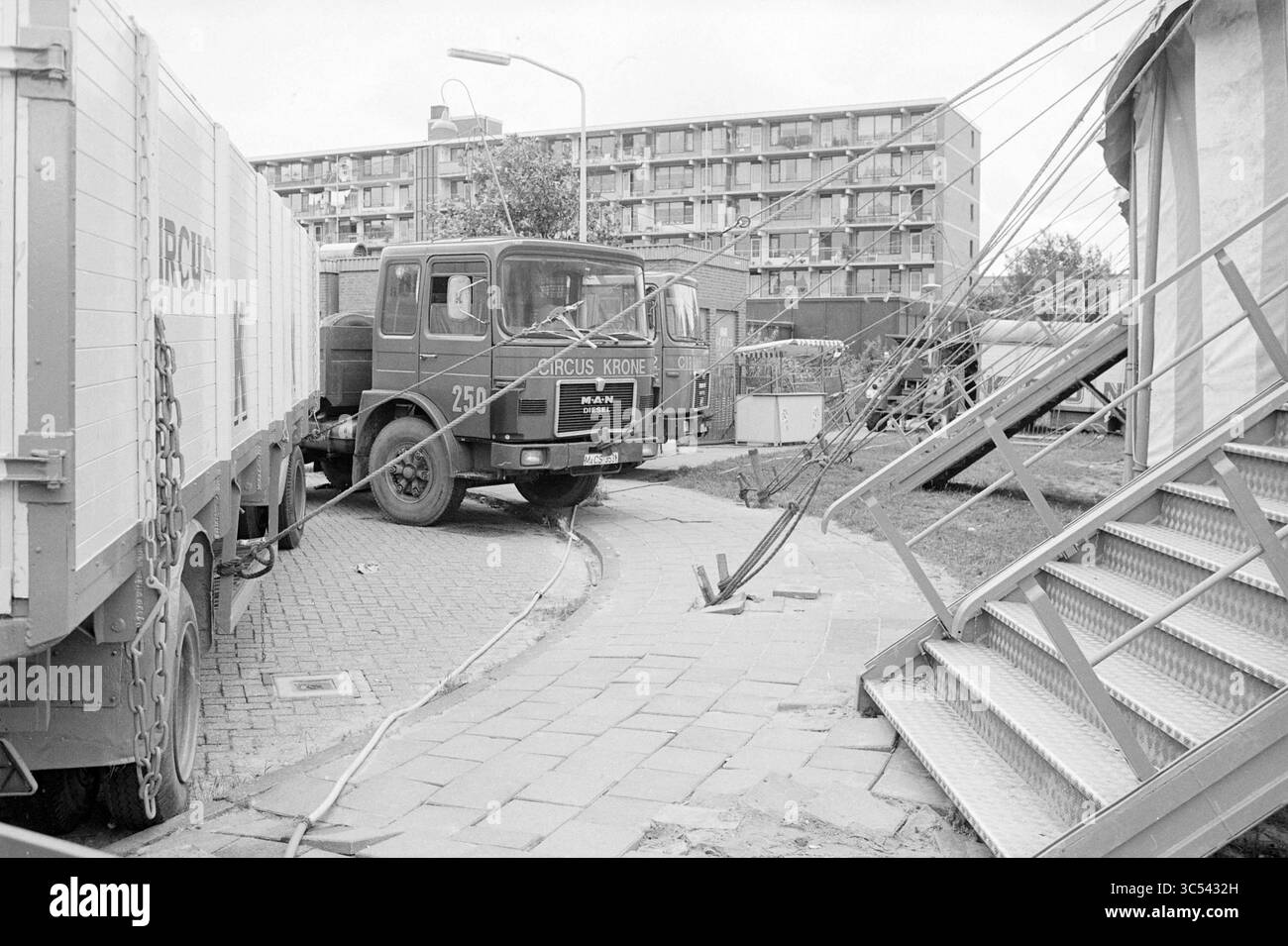 Wagons im Circus Krone mit Seilen, Circus, 05-08-1985 Whizgle News, Dutch Desk, Niederlande, 1950-2000 Eine Vintage-Zirkusszene mit einem großen Truck mit dem „Circus Kromer“, der in der Nähe einer Treppe zum Zelt geparkt ist. In der Nähe sind zusätzliche Lkw zu sehen, neben der Kulisse eines Wohngebäudes. Kabel sind vom Zelt entfernt, was die lebhafte Atmosphäre des Zirkus unterstreicht. Stockfoto