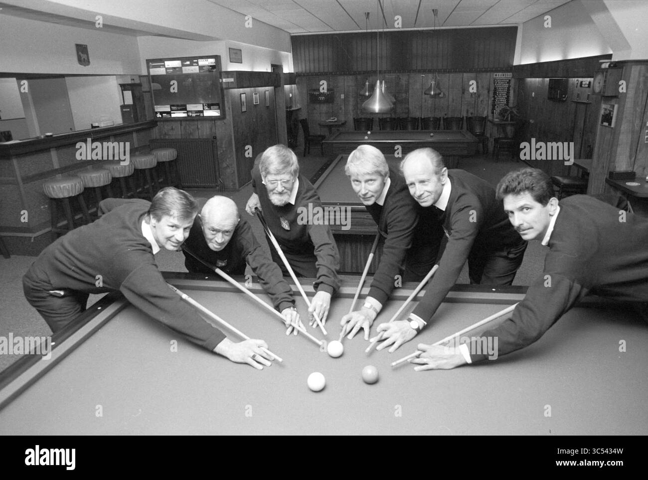 Billardclub, 00-00-1990 Whizgle News, Dutch Desk, Niederlande, 1950-2000 Eine Gruppe von sechs Personen, die in dunkler Kleidung gekleidet sind, versammeln sich um einen Billardtisch, jeder greift mit Hinweisen in der Hand auf die Bälle zu. Das Ambiente bietet ein klassisches Bar-Ambiente mit Holztäfelung und Hockern im Hintergrund. Ihre Ausdrucksformen deuten auf Kameradschaft und Konzentration hin, während sie sich auf ein Spiel vorbereiten. Stockfoto