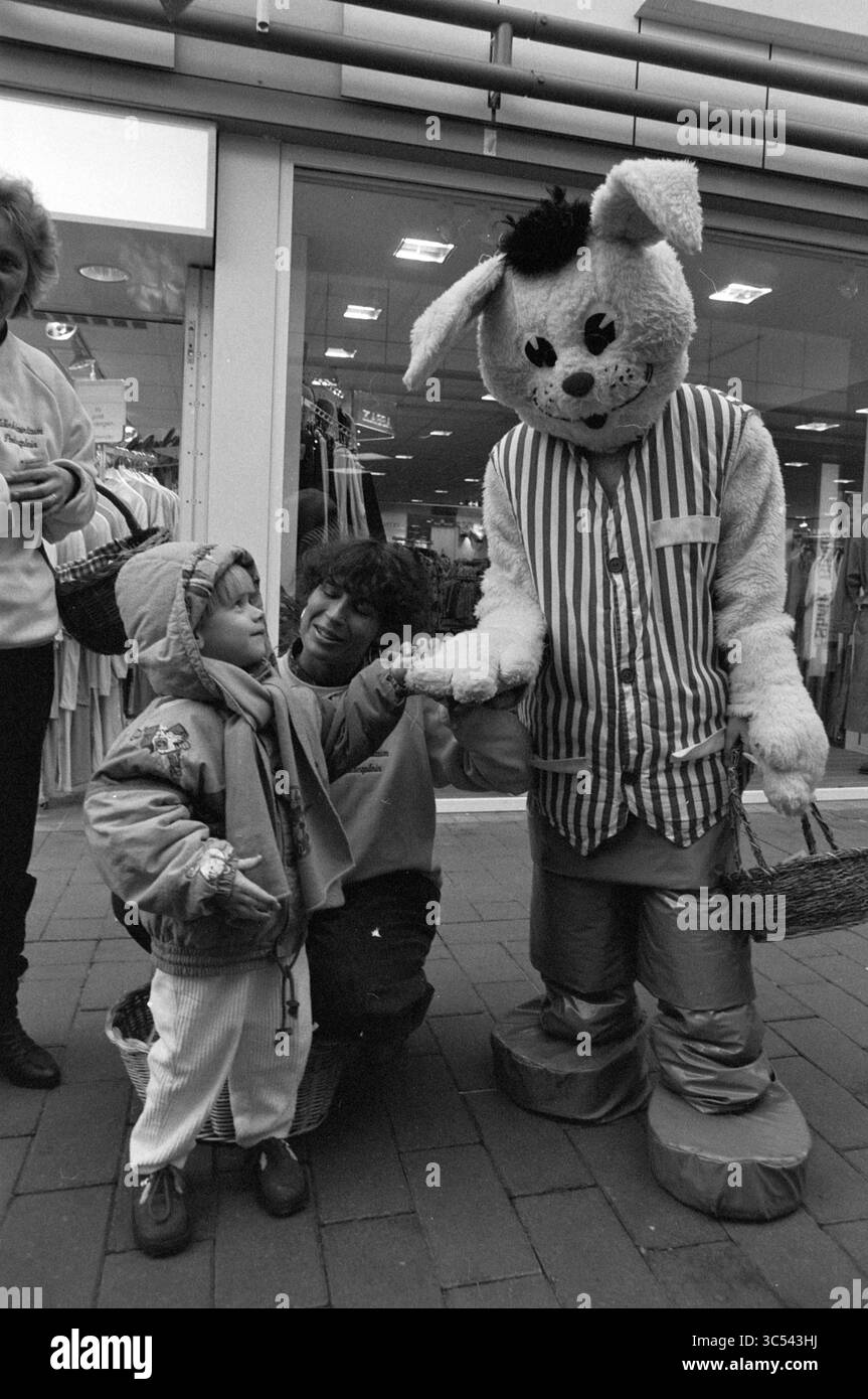 Valentinstag Kampagne Polderplein Hoofddorp, Valentinstag, Hoofddorp, Polderplein, Niederlande, 14-02-1990 Whizgle News, Dutch Desk, Niederlande, 1950-2000 Ein Kind in einem gemütlichen Outfit interagiert mit einer Person, die als Kaninchen gekleidet ist, die ein gestreiftes Outfit trägt und einen Korb hält. In der Nähe sieht ein Erwachsener mit einem Lächeln zu und schafft eine verspielte Szene voller Freude und Neugier. Stockfoto