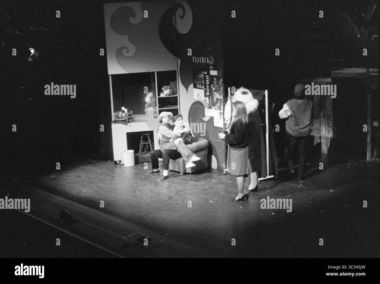 Musical Stadsschouwburg IJmuiden, IJmuiden, Niederlande, 12-05-1990 Whizgle News, Dutch Desk, Niederlande, 1950-2000 Eine lebendige Szene aus einer Theateraufführung, in der vier Charaktere verschiedene Aktivitäten ausüben: zwei sitzen auf einem Sofa, eine andere steht in der Nähe einer Trennwand und eine bereitet sich seitlich vor. Das Set verfügt über eine stilisierte Kulisse mit dekorativen Elementen, die die dramatische Atmosphäre unterstreichen. Stockfoto