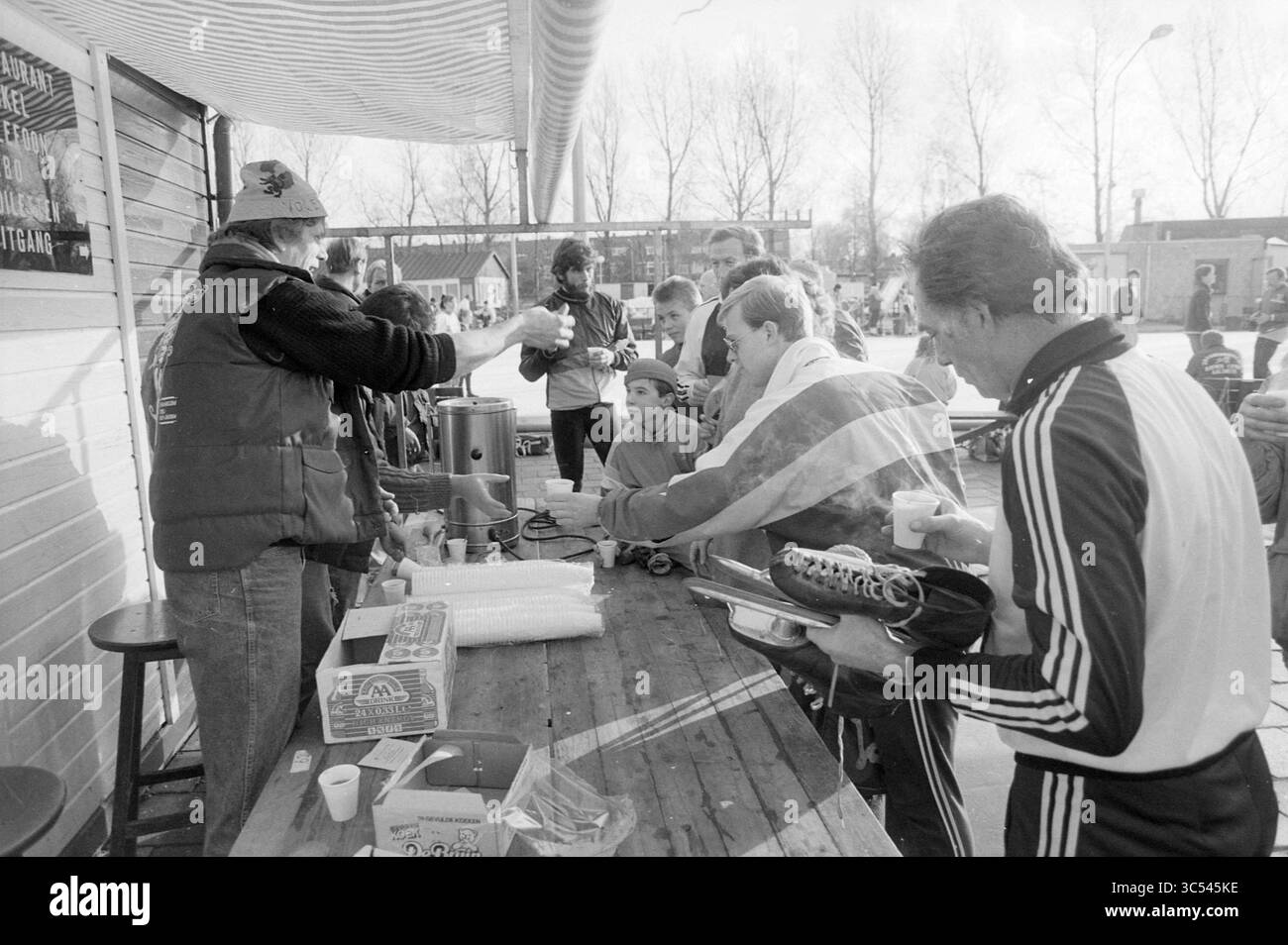 Koek en zopie Eislaufbahn gratis Kokos, Eis, Eislaufbahn, Eishockey, Haarlem, IJsbaanlaan, Niederlande, 27-11-1988 Whizgle News, Dutch Desk, Niederlande, 1950-2000 Eine Gruppe von Menschen versammelt sich an einem Holztisch und genießt warme Getränke und Snacks. Einige tragen Wintersportausrüstung, während andere sich lebhaft unterhalten. Ein Anbieter serviert Getränke aus einem großen Behälter, wodurch ein Gefühl von Gemeinschaft und Kameradschaft unter den Teilnehmern entsteht. Stockfoto
