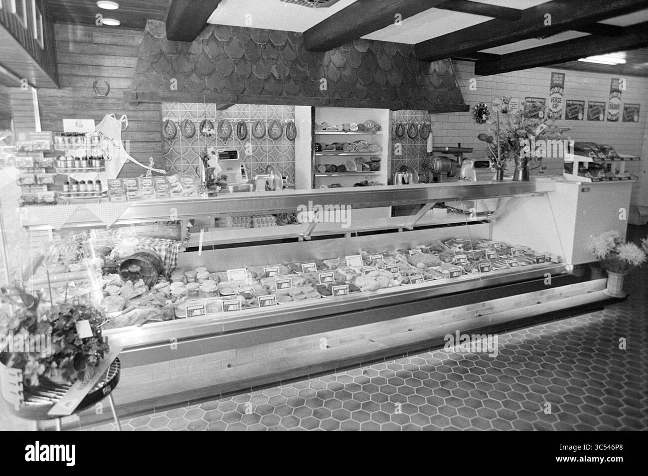 Intr. Fa. v Eyk Slagerij Heemstede, Interior, 17-07-1973 Whizgle News, Dutch Desk, Niederlande, 1950-2000 Eine gut organisierte Ausstellung zeigt eine Vielzahl von Fleisch- und Käsesorten und verpackten Waren, die alle ordentlich hinter einer Glastheke angeordnet sind. Das rustikale Dekor mit freiliegenden Balken und Fliesenakzenten verleiht dem Markt eine gemütliche Atmosphäre. Frische Blumen und Dekorationsartikel unterstreichen das einladende Ambiente. Stockfoto