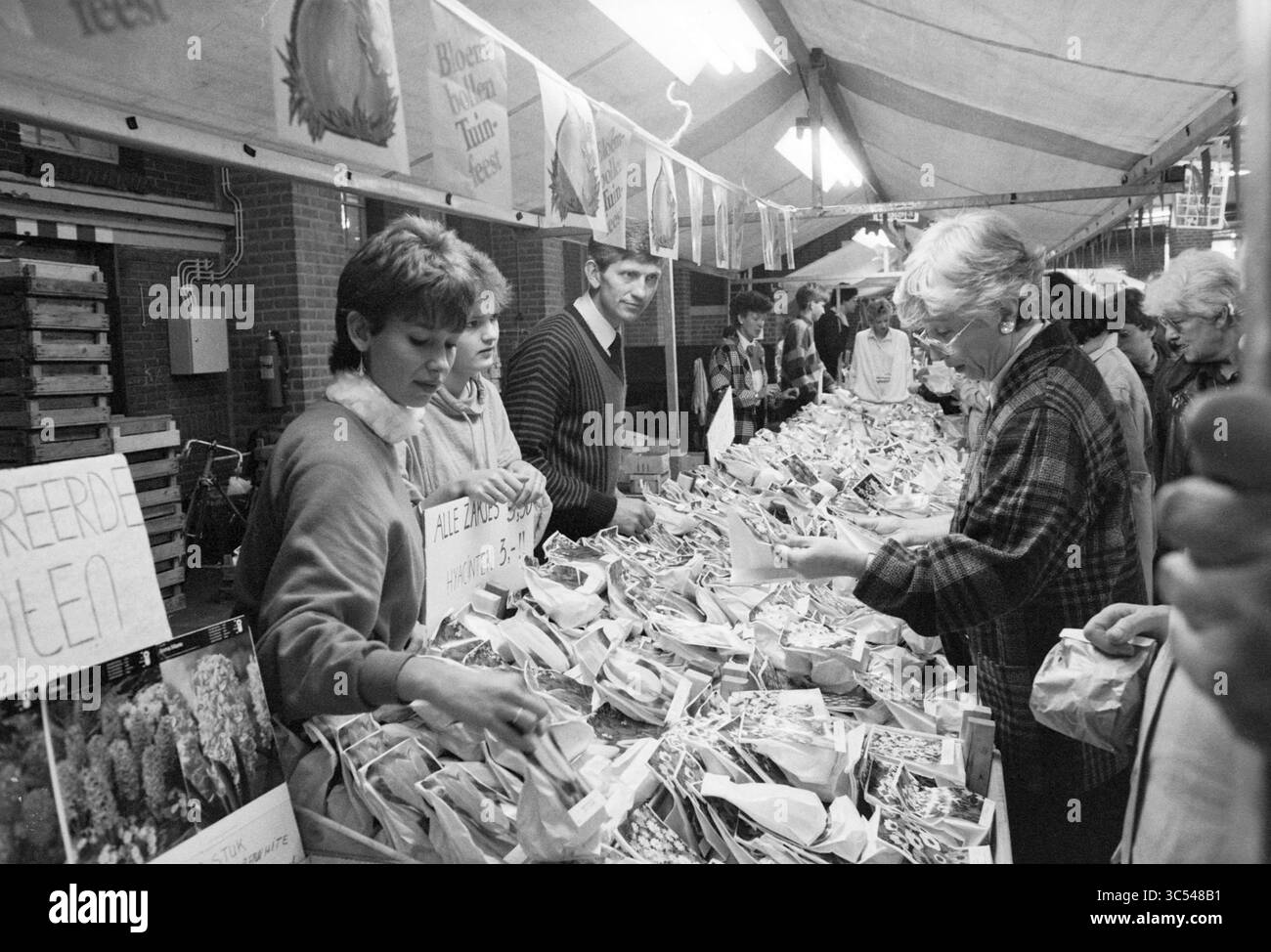 Nass. Blumenzwiebelmarkt, Blumenzwiebeln, 19-10-1985 Whizgle News, Dutch Desk, Niederlande, 1950-2000 Eine geschäftige Marktszene voller Menschen, die an verschiedenen Verkaufsständen interagieren, an denen Anbieter eine Vielzahl von verpackten Waren anbieten. Einige wählen sorgfältig Elemente aus, während andere chatten, um eine lebhafte Atmosphäre zu schaffen. Banner und Schilder weisen auf die verfügbaren lokalen Produkte hin und tragen zur lebendigen Interaktion mit der Community bei. Stockfoto