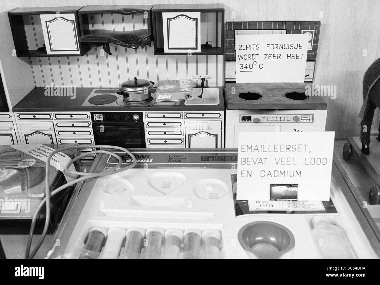 Herr Kippersluis Inspektionsdienst von Waren, Inspektionen, Personen, 08-10-1981 Whizgle News, Dutch Desk, Niederlande, 1950-2000 Eine Miniatur-Kücheneinrichtung mit einem Zweiflammer-Herd mit einem Warnschild für hohe Temperaturen sowie einer Spielküche mit Backofen. Verschiedene Laborgeräte, einschließlich Reagenzgläser und Behälter, sind im Vordergrund angeordnet und mit Sicherheitsinformationen zu Gefahrstoffen gekennzeichnet. Stockfoto