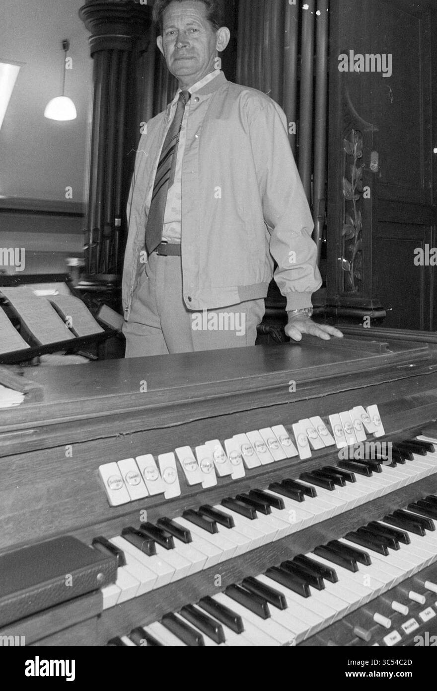 Orgelkirche HaarlemmerLiede, 24-08-1982 Whizgle News, Dutch Desk, Niederlande, 1950-2000 Ein Mann steht selbstbewusst neben einer Vintage-Orgel, in formeller Kleidung mit gemusterter Krawatte gekleidet. Die Tasten der Orgel, die teilweise mit einzigartigen Symbolen gekennzeichnet sind, deuten auf die im Raum gespielte Musik hin, während verzierte Pfeifen im Hintergrund aufsteigen. Stockfoto