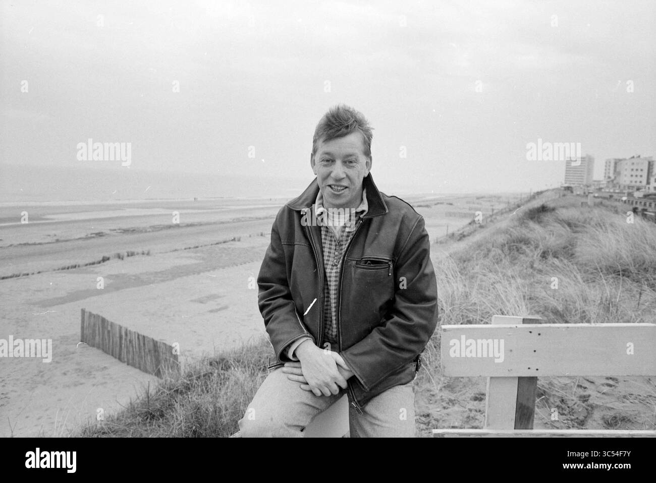Jelle de Jong, Z'voort, Zandvoort, 21.01.1997 Whizgle News, Dutch Desk, Niederlande, 1950-2000 Ein Mann sitzt lässig auf einer Bank mit Blick auf einen Sandstrand, mit Blick auf das Meer im Hintergrund und Gebäude am Ufer. Er trägt eine Jacke und lächelt, während er entspannt in einem bewölkten Himmel erscheint. Stockfoto