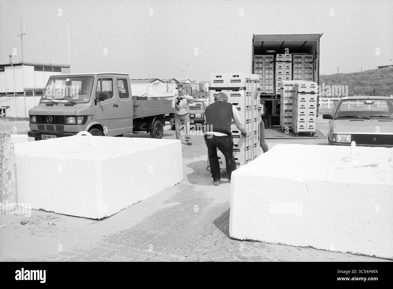 LKW-Entladegeschäfte IJmuiden Beach, Geschäfte und Kioske, IJmuiden, Niederlande, 11-05-1989 Whizgle News, Dutch Desk, Niederlande, 1950-2000 Ein Mann schiebt einen Wagen auf einen mit Kisten beladenen LKW, während eine andere Person in der Nähe in einer belebten Umgebung steht. Die Szene zeigt große, weiße Betonblöcke auf dem Boden, die auf einen belebten Ladebereich hinweisen. Im Hintergrund ist eine Mischung aus Fahrzeugen und Strukturen zu sehen, die auf ein gewerbliches oder industrielles Umfeld hinweist. Stockfoto